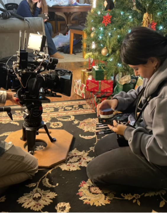 Person sitting on a patterned rug near a decorated Christmas tree with presents, holding a phone, with a camera on a tripod nearby.