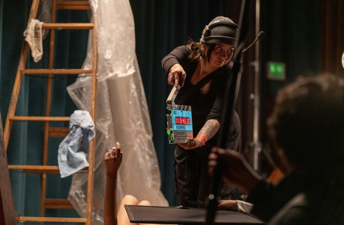 Person on stage holding a small object, with a ladder and fabric in the background, and audience members nearby.