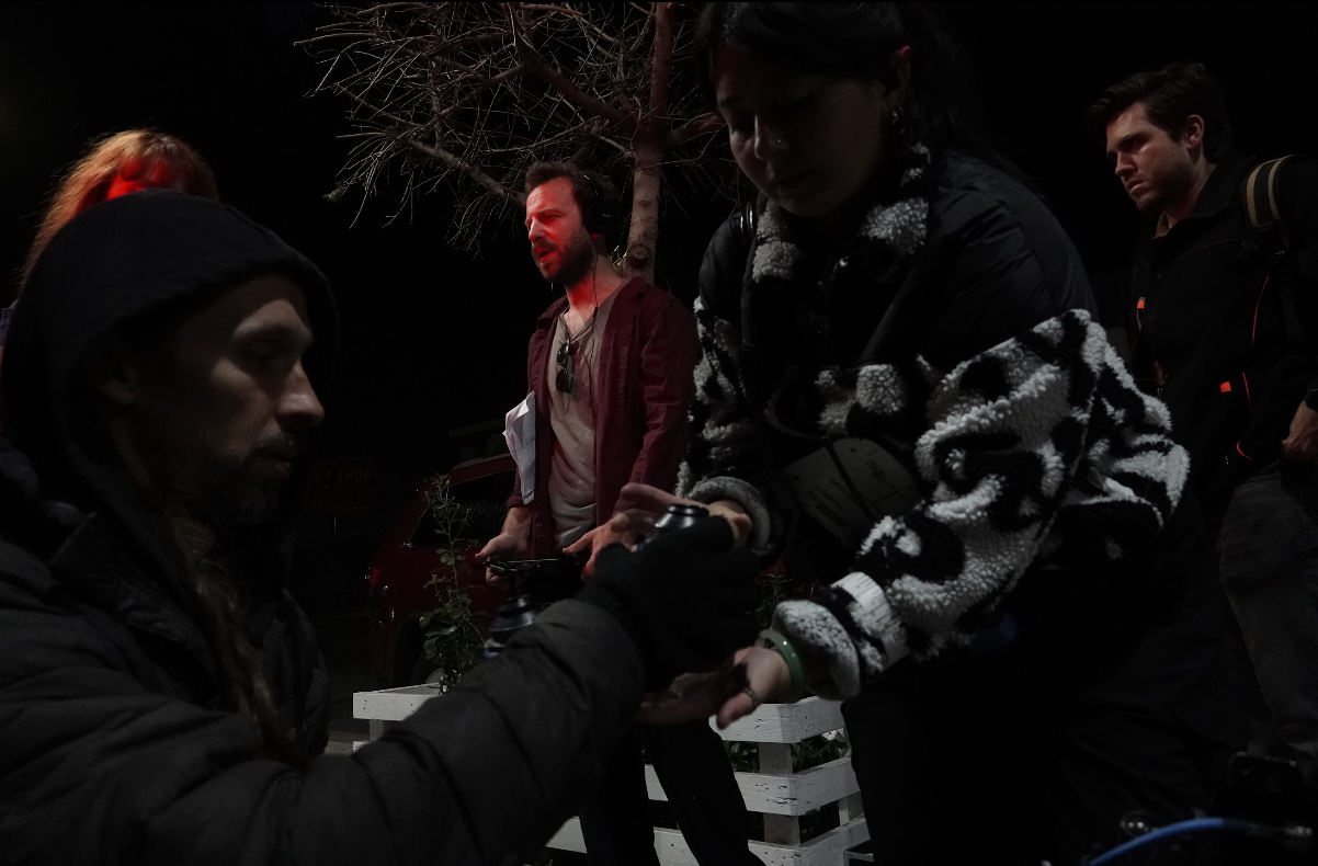 Group of people in a dark setting, some interacting, with one person handing something to another, and others in the background.