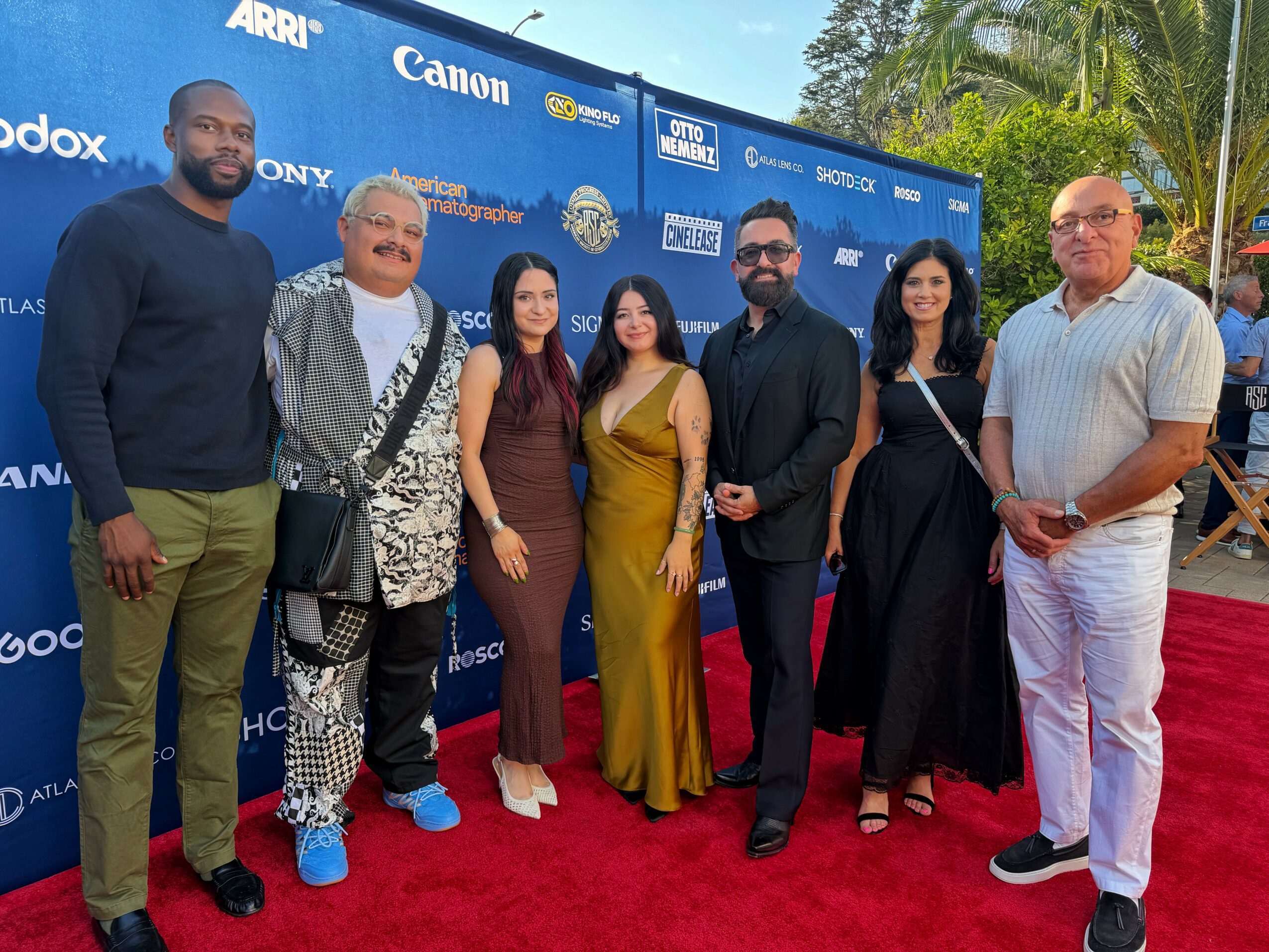 Group of seven people standing on a red carpet outdoors, smiling, with a blue backdrop featuring logos behind them.