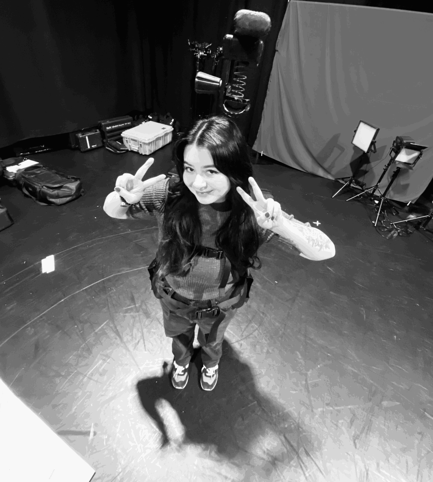 Young woman with long hair making peace signs with both hands, smiling, standing on a stage with equipment around.