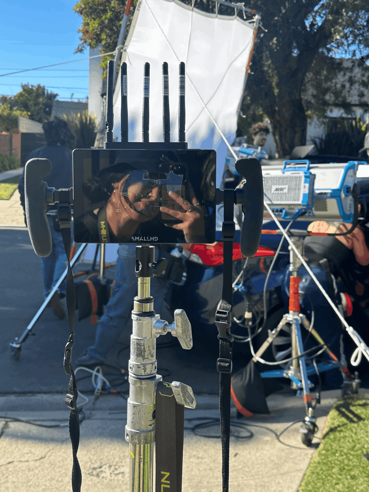 Outdoor scene with camera setup and people, trees, and chairs in background.