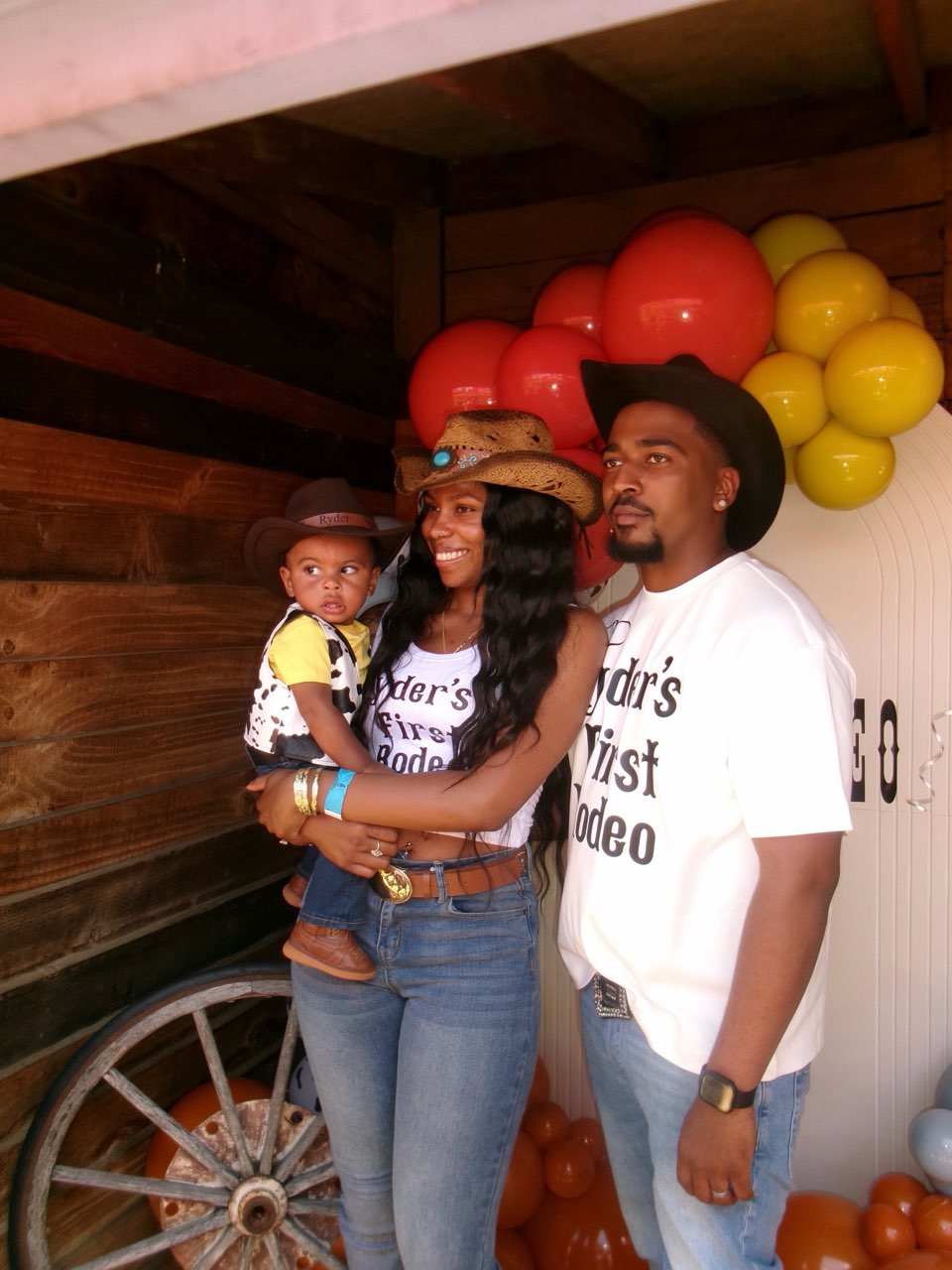 Three people, a woman holding a child and a man, stand together in front of balloons and wooden background.