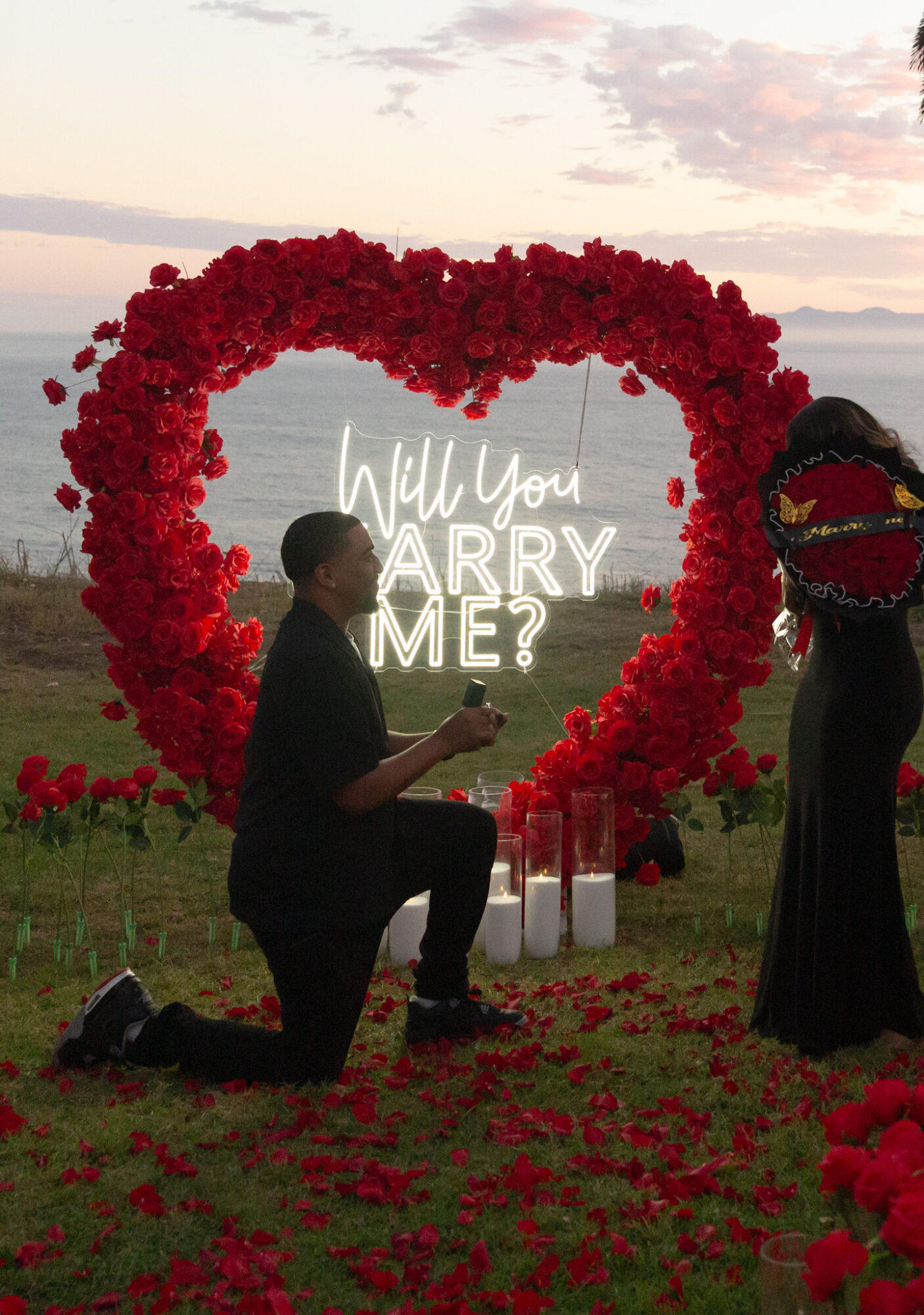 Person kneeling with a ring in front of a heart-shaped flower arch and a neon sign that says