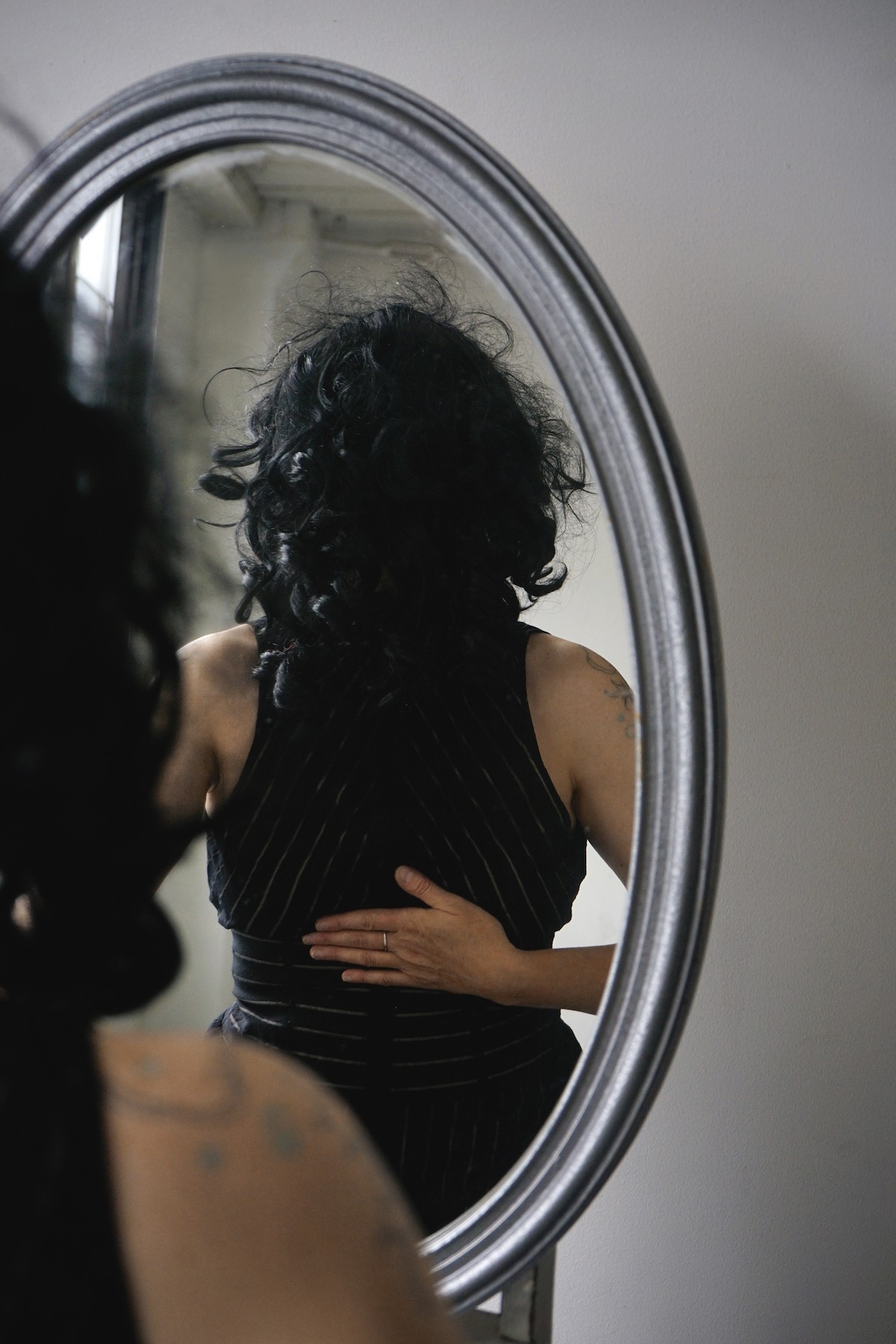 Person with curly hair looking into an oval mirror, hand on their stomach, wearing a dark striped top.