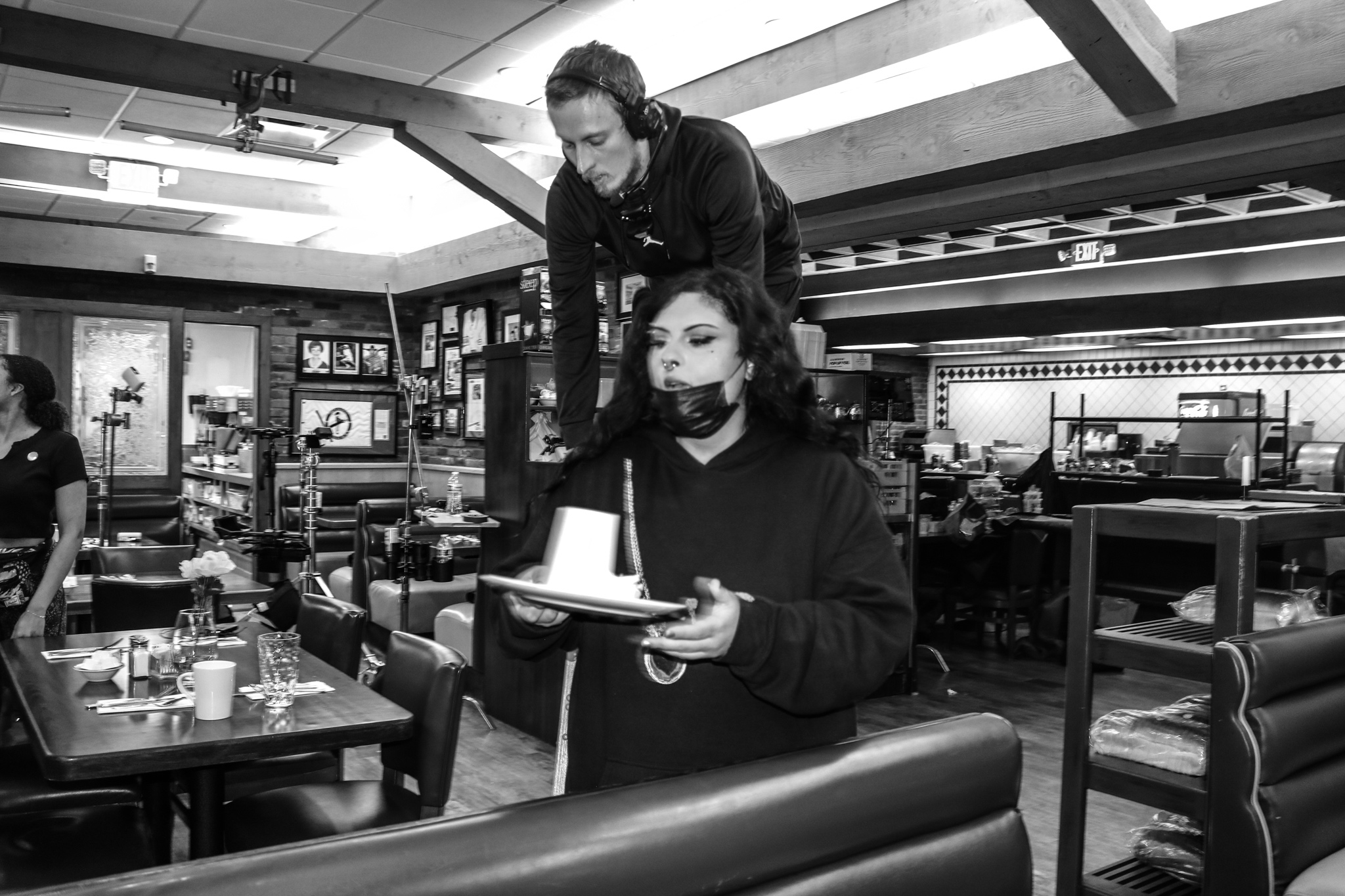 Two women in a restaurant, one standing and the other sitting, with tables and shelves in the background.