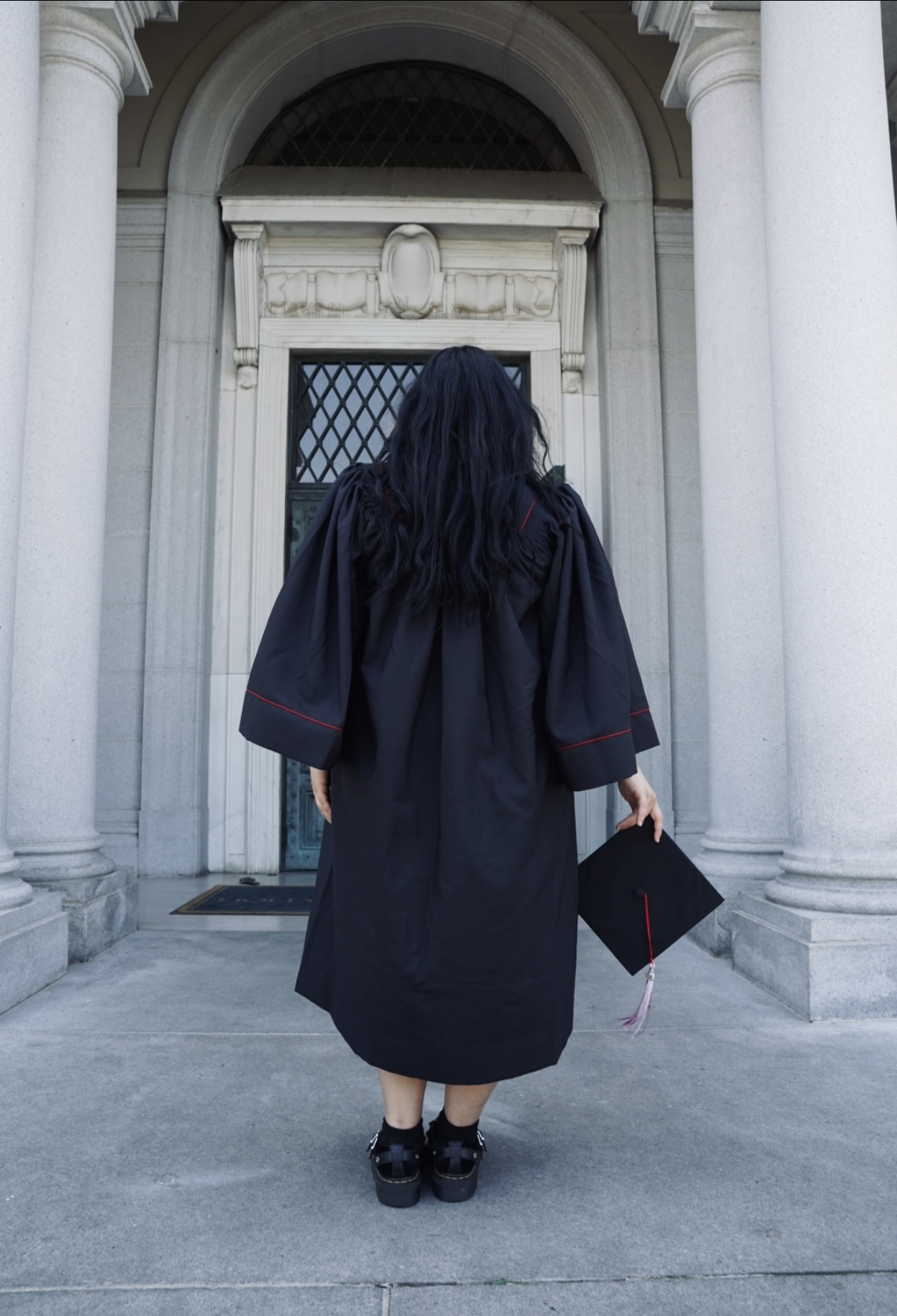 Person in a graduation gown standing in front of a building with columns and an arched entrance, holding a diploma.