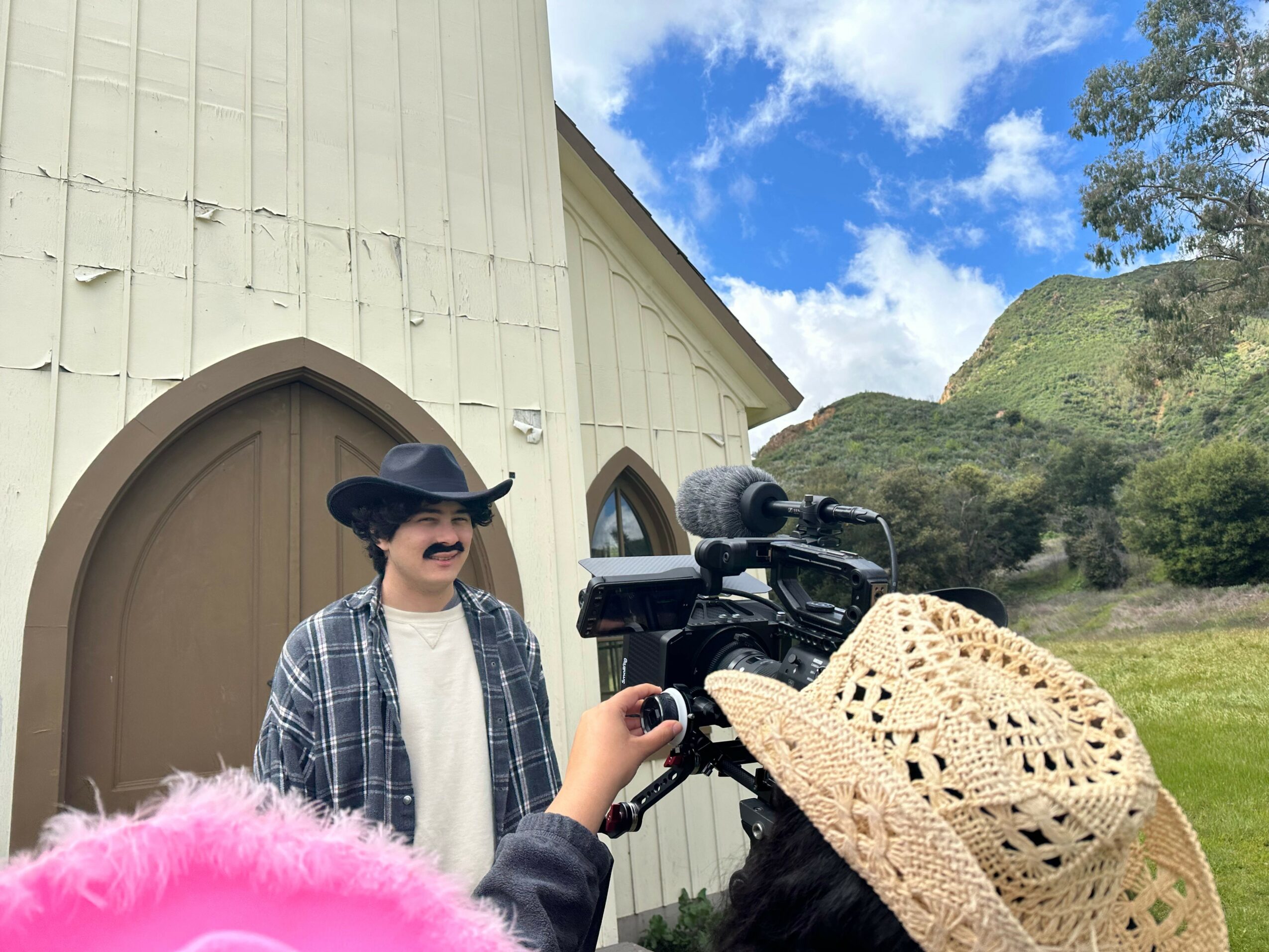 Person wearing a straw hat and dark jacket operating a camera outdoors, with a white building and green hills in the background.
