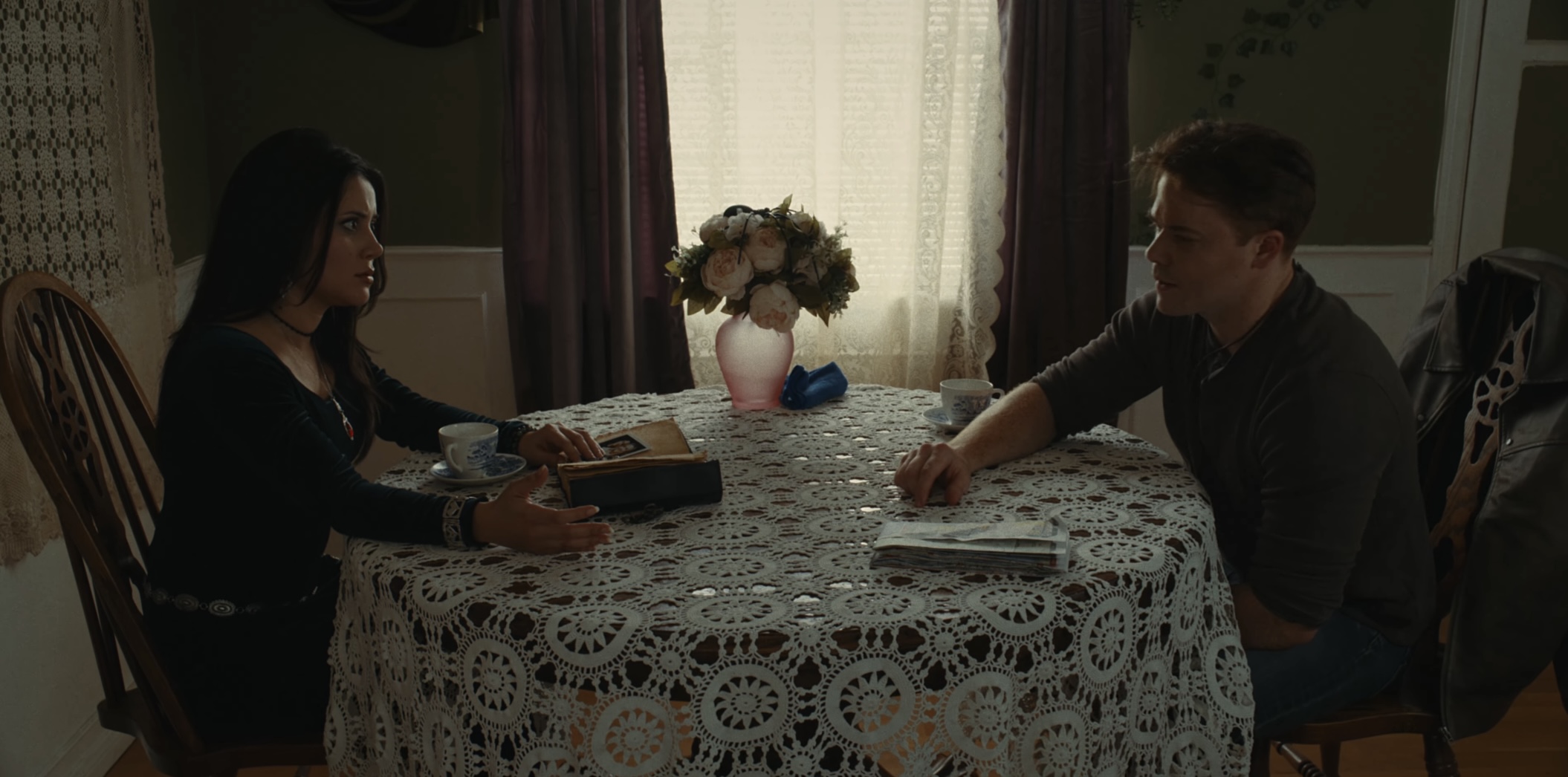 Two people sit at a table with a lace tablecloth, a flower vase, and books, in a room with curtains.