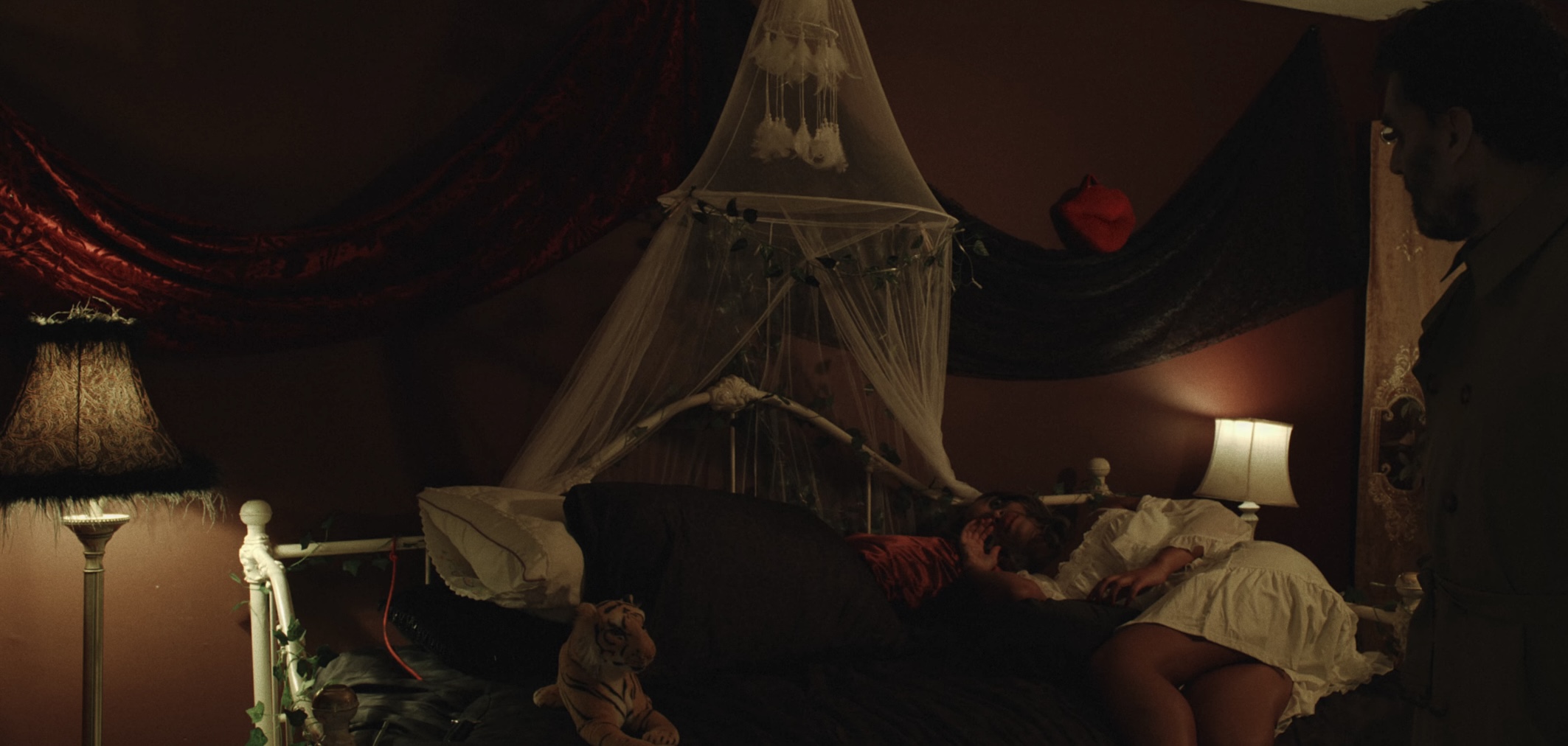 Person lying on bed with a canopy, in a dimly lit room with lamps and dark curtains.