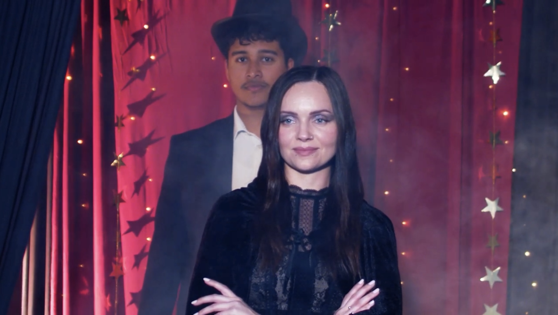 Two people standing in front of a decorated backdrop with stars and lights, one woman in front and one man behind.