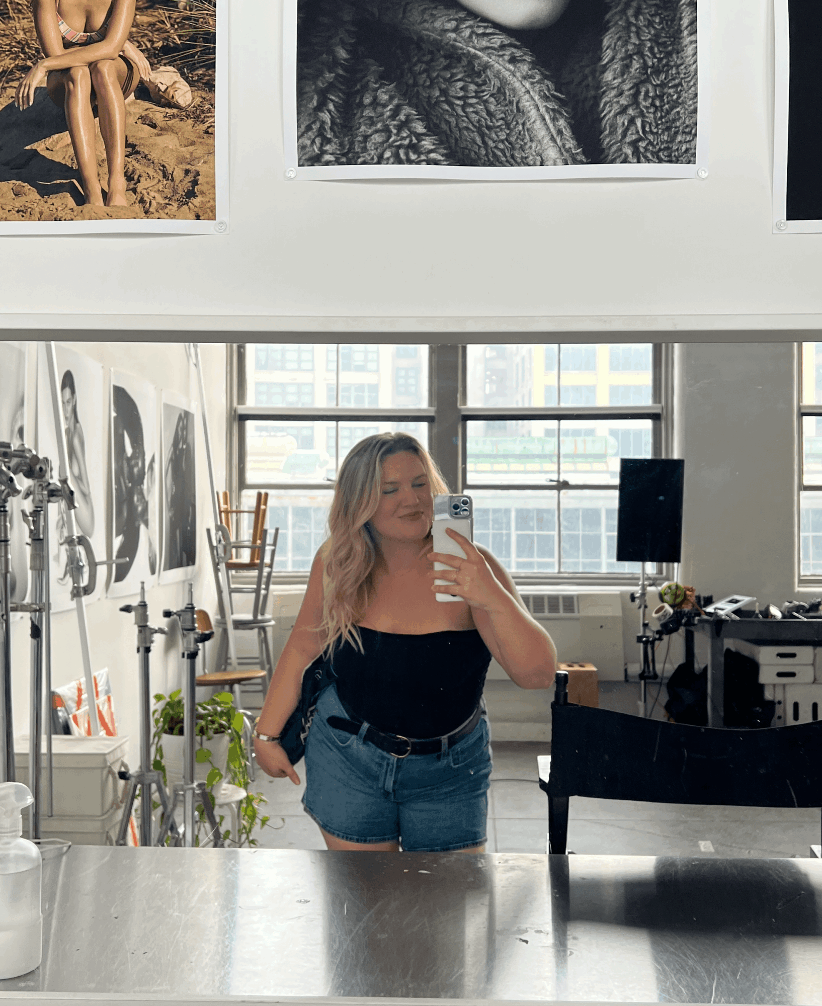 Woman taking a mirror selfie in a bright room with large windows, art supplies, and plants, wearing a black top and denim shorts.