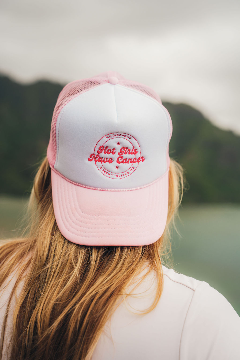 Person with long hair wearing a pink and white cap outdoors, with a cloudy sky and mountains in the background.