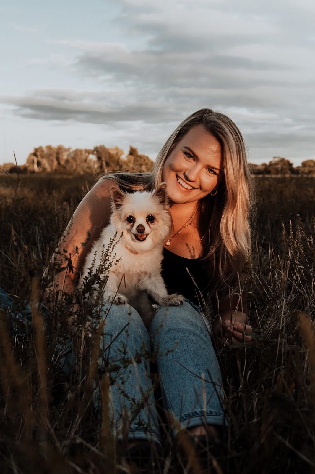 Woman smiling with a small dog in a grassy field during sunset, cloudy sky in background.