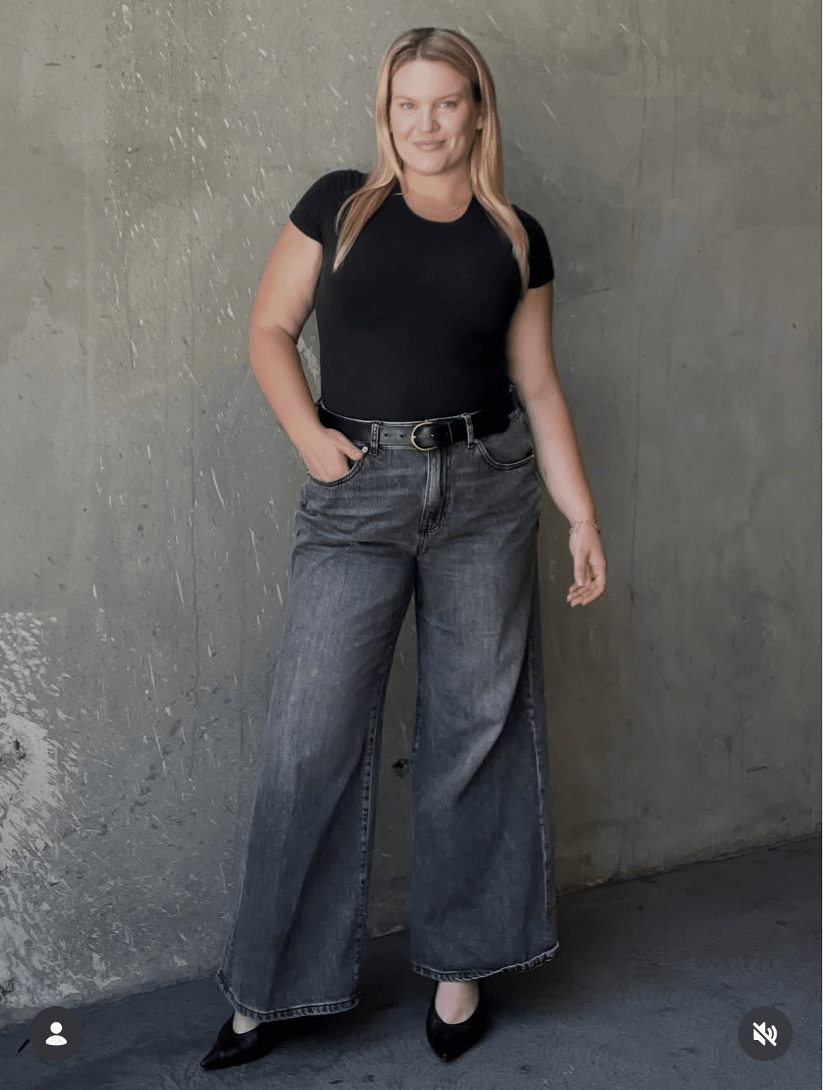 Woman standing against a concrete wall, wearing a black t-shirt, wide-leg jeans, and black shoes, with one hand in her pocket.