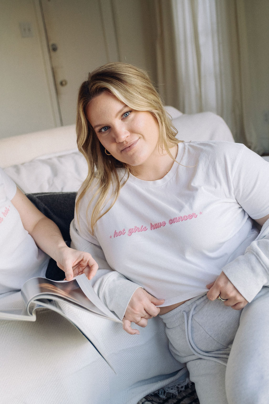 Young woman with blonde hair sitting on a couch, smiling, wearing a white shirt and light-colored pants, holding a magazine.