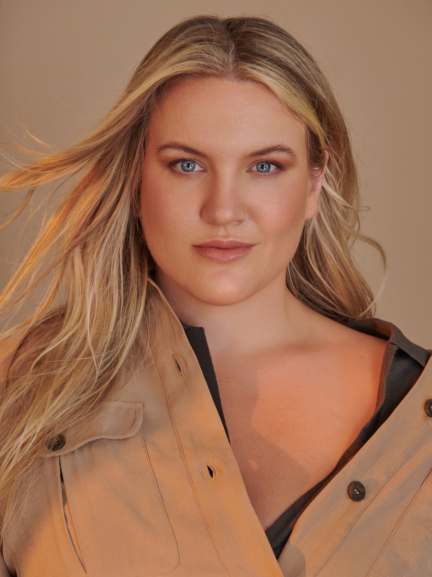 Portrait of a young woman with long blonde hair and blue eyes, wearing a beige jacket, looking directly at the camera.