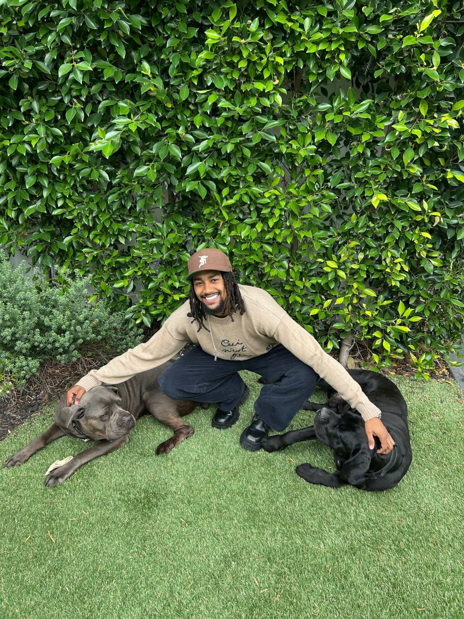 Person with glasses and beige sweater lying on grass with two dogs, one black and one gray, near bushes.