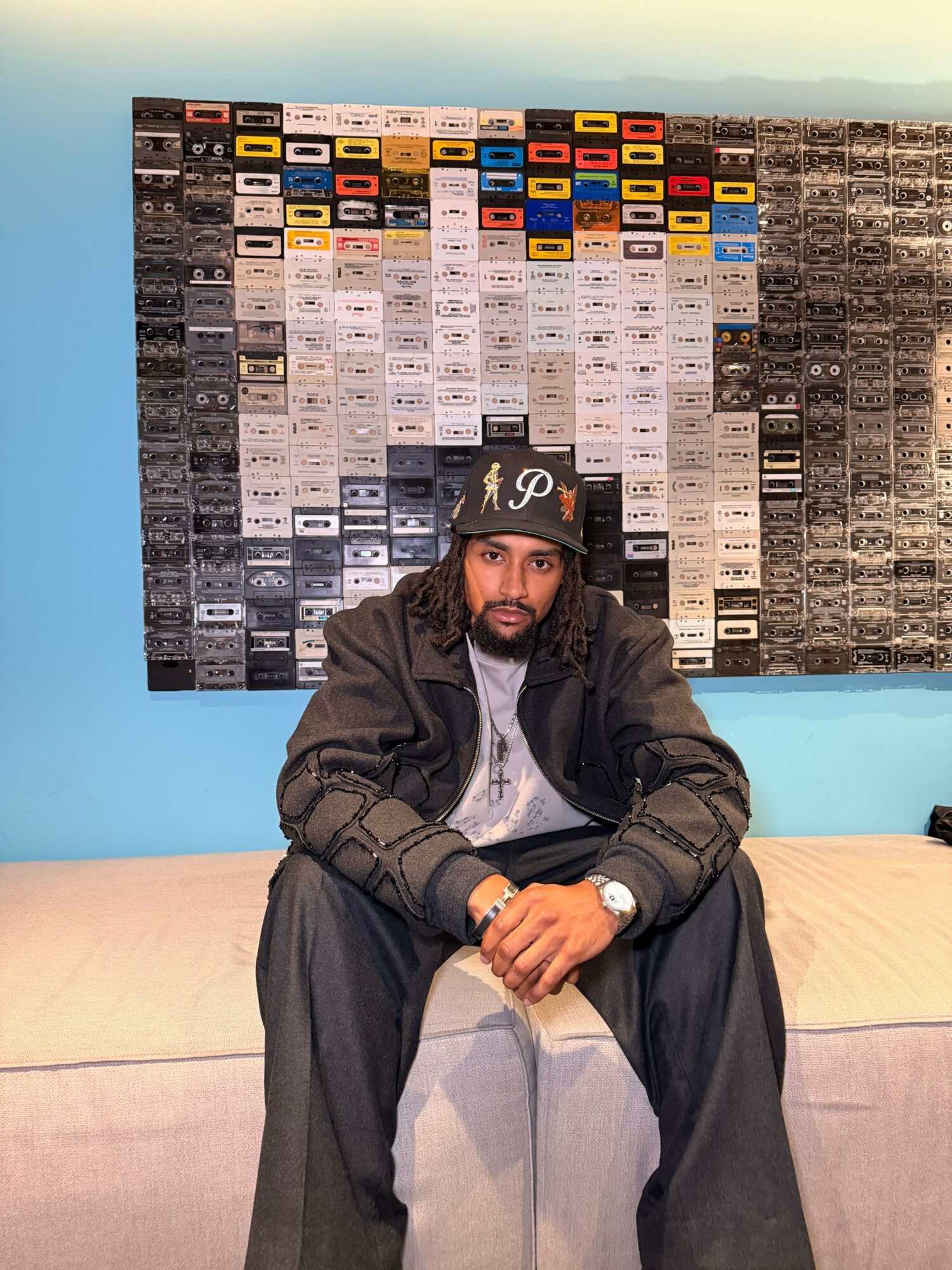 Person wearing a black jacket and cap, sitting on a pink couch, with a colorful wall of cassette tapes behind them.