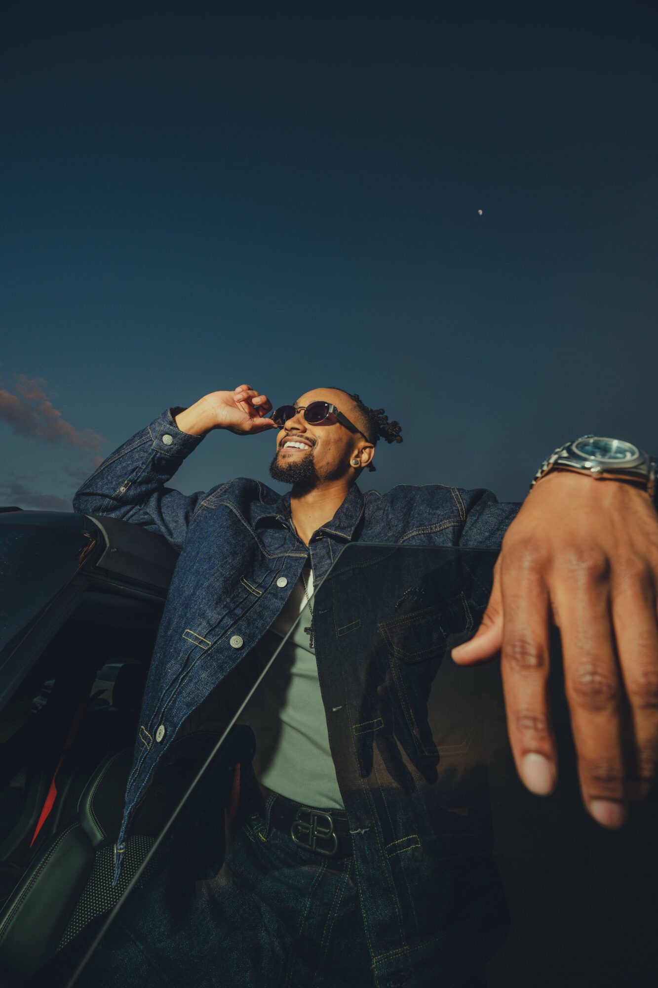 Person with sunglasses smiling, wearing a denim jacket, leaning on a car against a dark sky with a star visible.