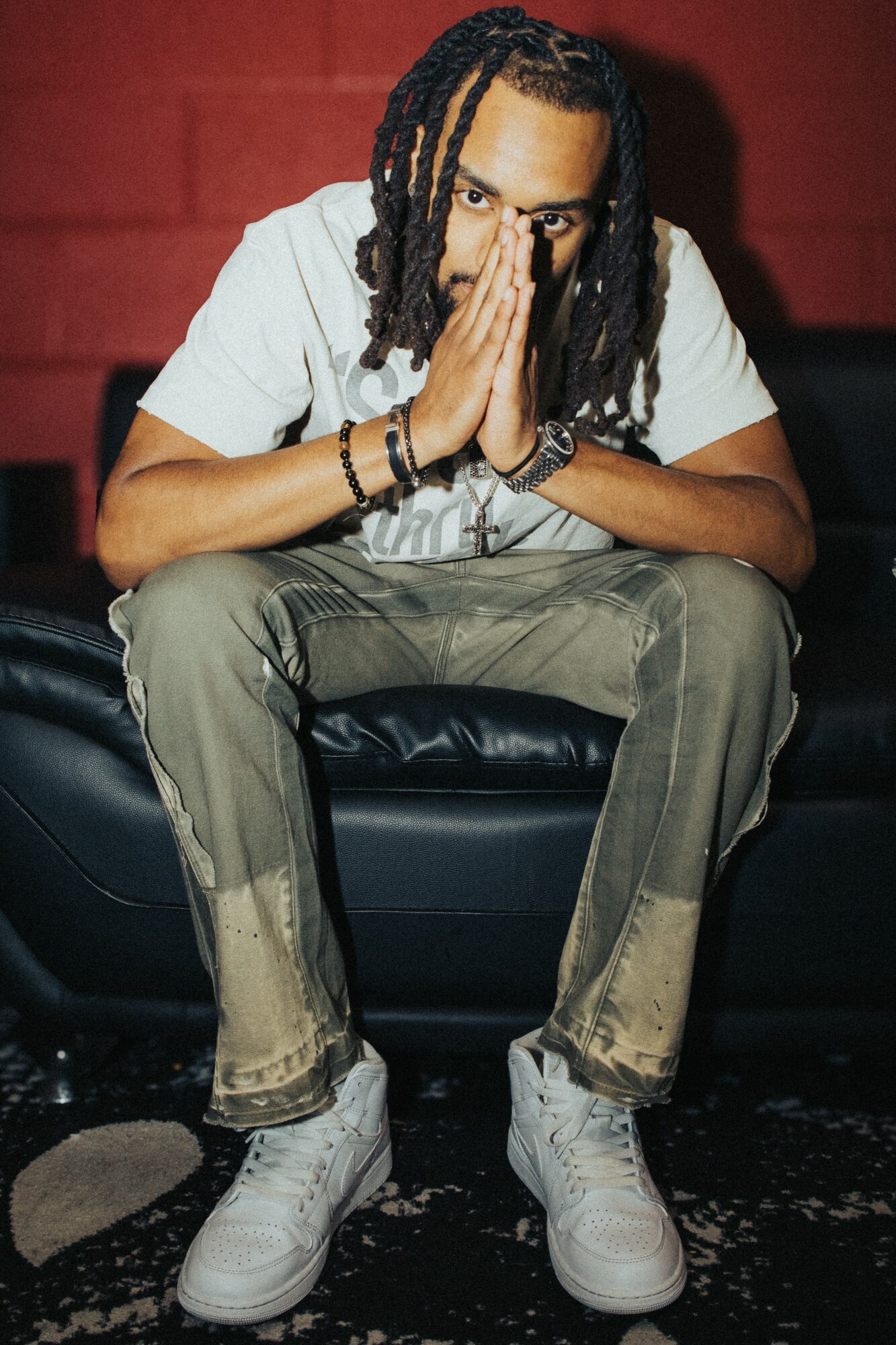 Person with dreadlocks sitting on a black couch, hands pressed together near face, wearing a white shirt and light-colored pants