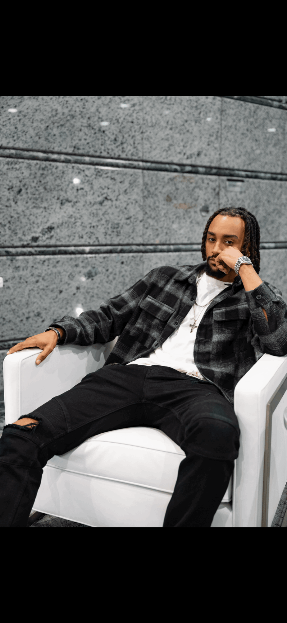 Person sitting on a white chair against a gray wall, wearing a black and white checkered shirt and black pants.