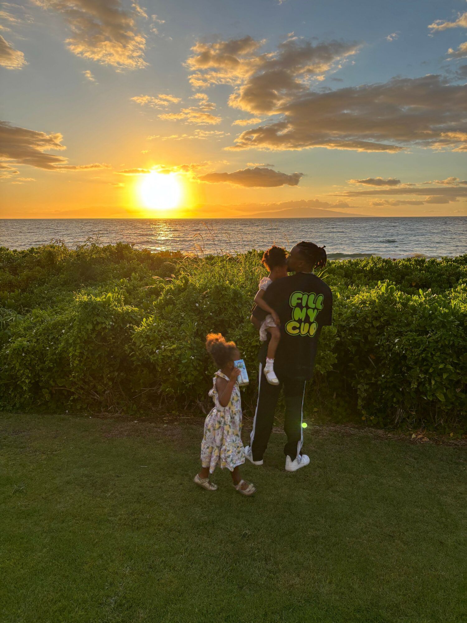 Two children standing on grass during sunset, with trees and a cloudy sky in the background.