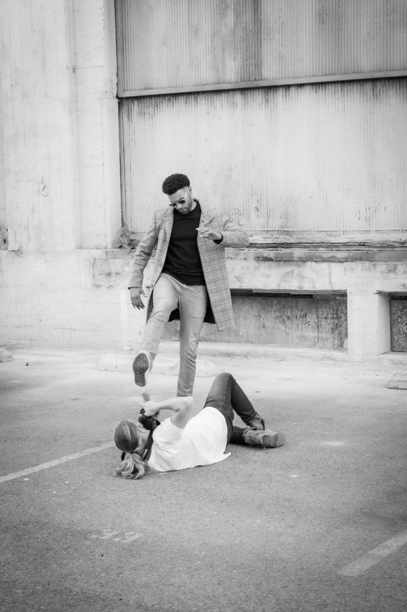 Two people on the ground in an urban setting, one standing and the other lying down, with concrete walls in the background.