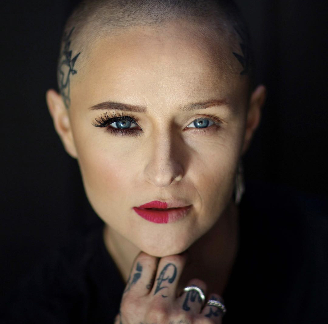 Close-up of a woman with short hair, tattoos, and makeup, looking directly at the camera with one eye slightly squinted.