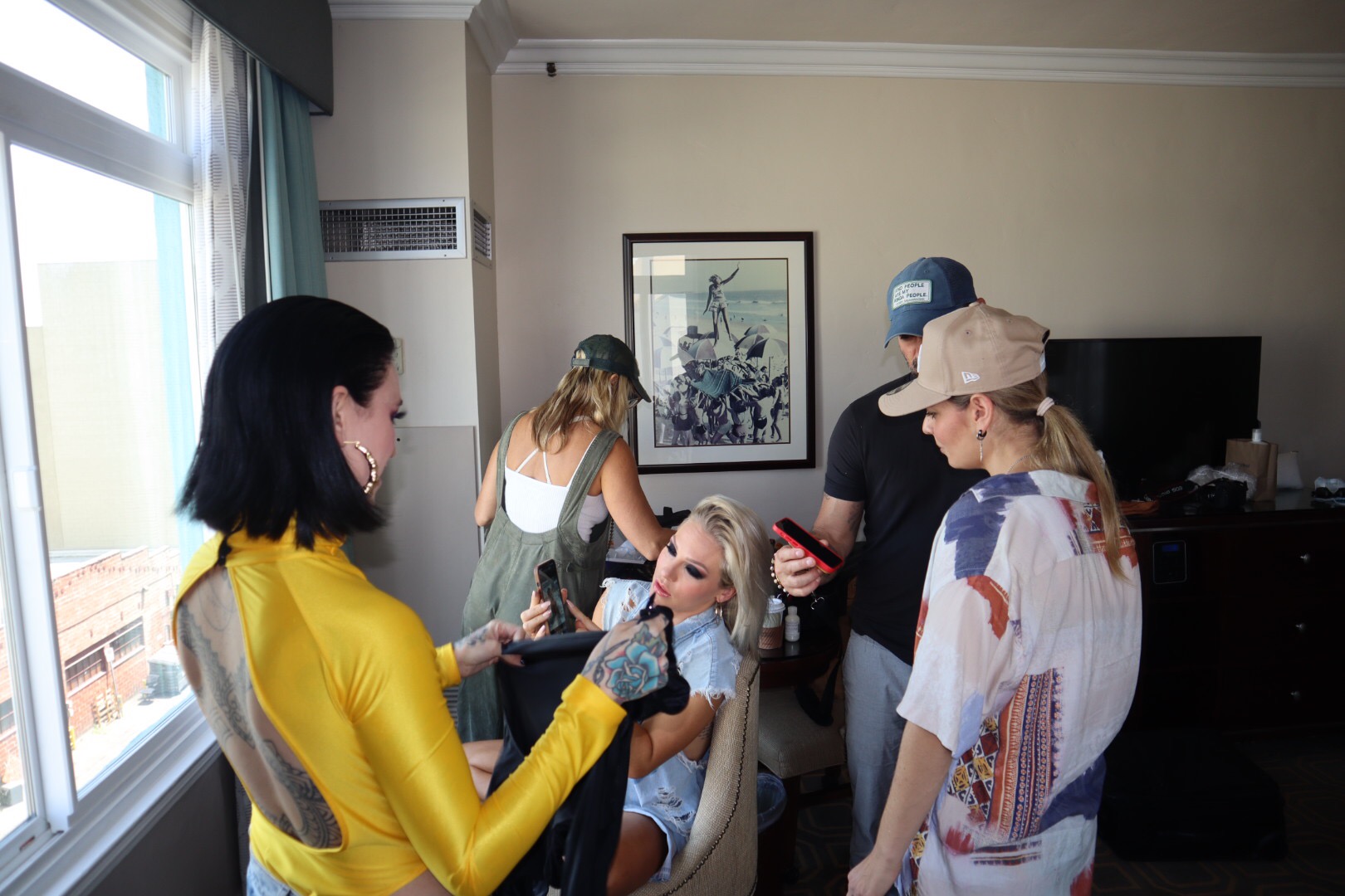 Group of five people in a room, one woman in yellow holding a phone, others gathered around, with a window and framed picture on the wall.