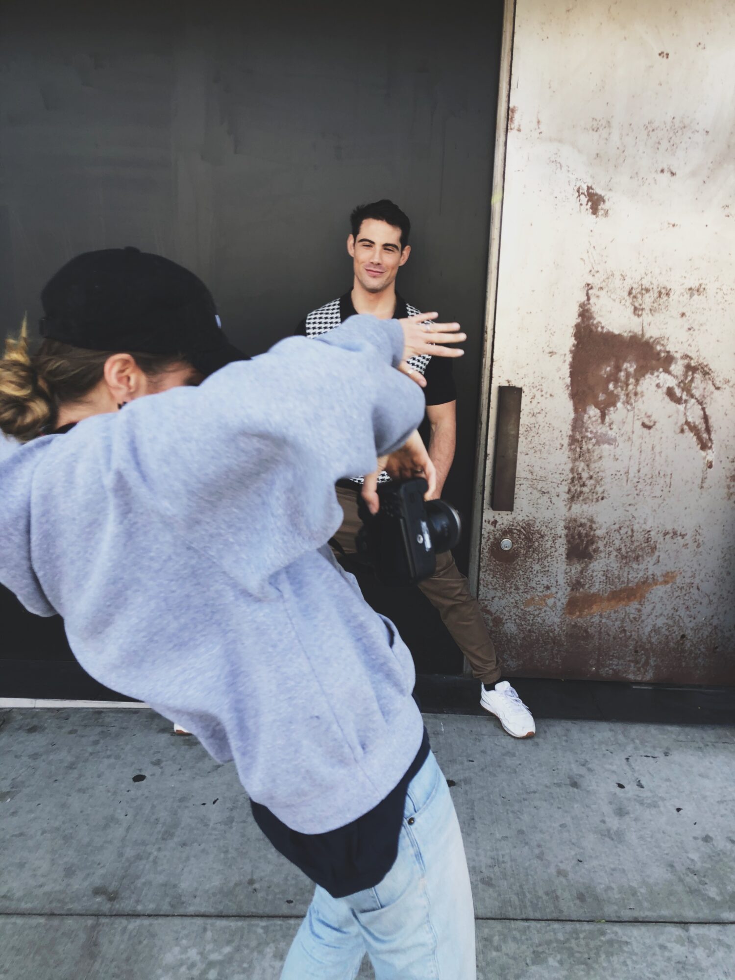 Person in a gray hoodie and light jeans gestures towards a young man sitting against a wall, with a black and white background.