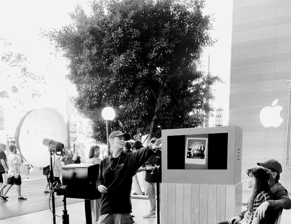People gather outside an Apple store with a large tree and a display screen, some taking photos or talking.
