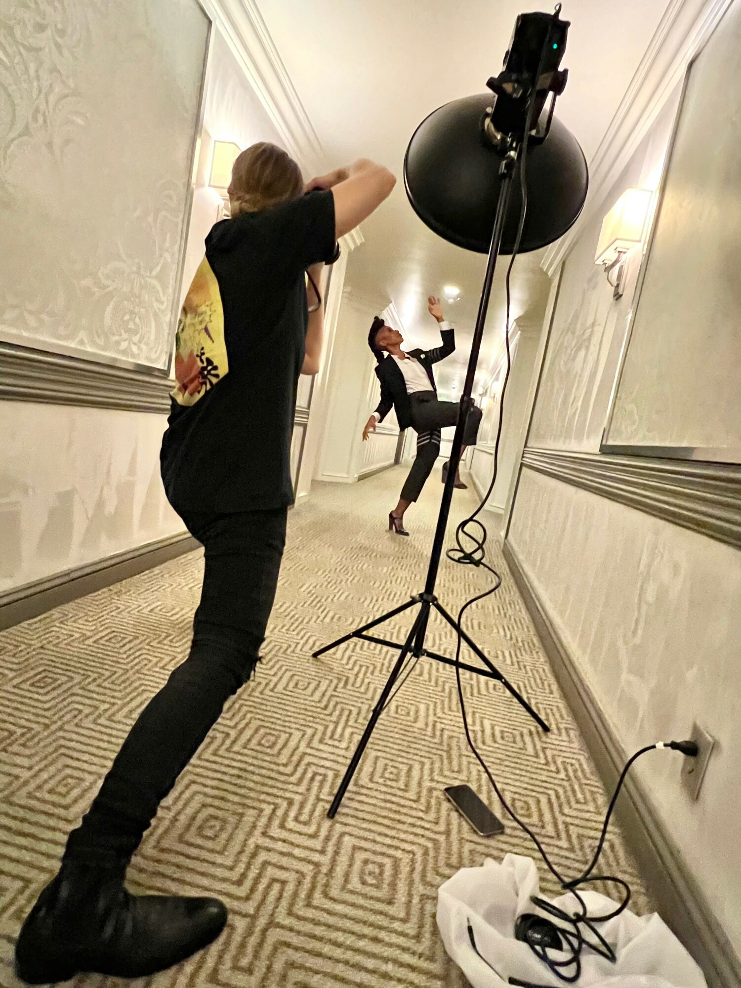 Two people in a hallway, one taking a photo and the other posing with a raised leg, with photography equipment nearby.