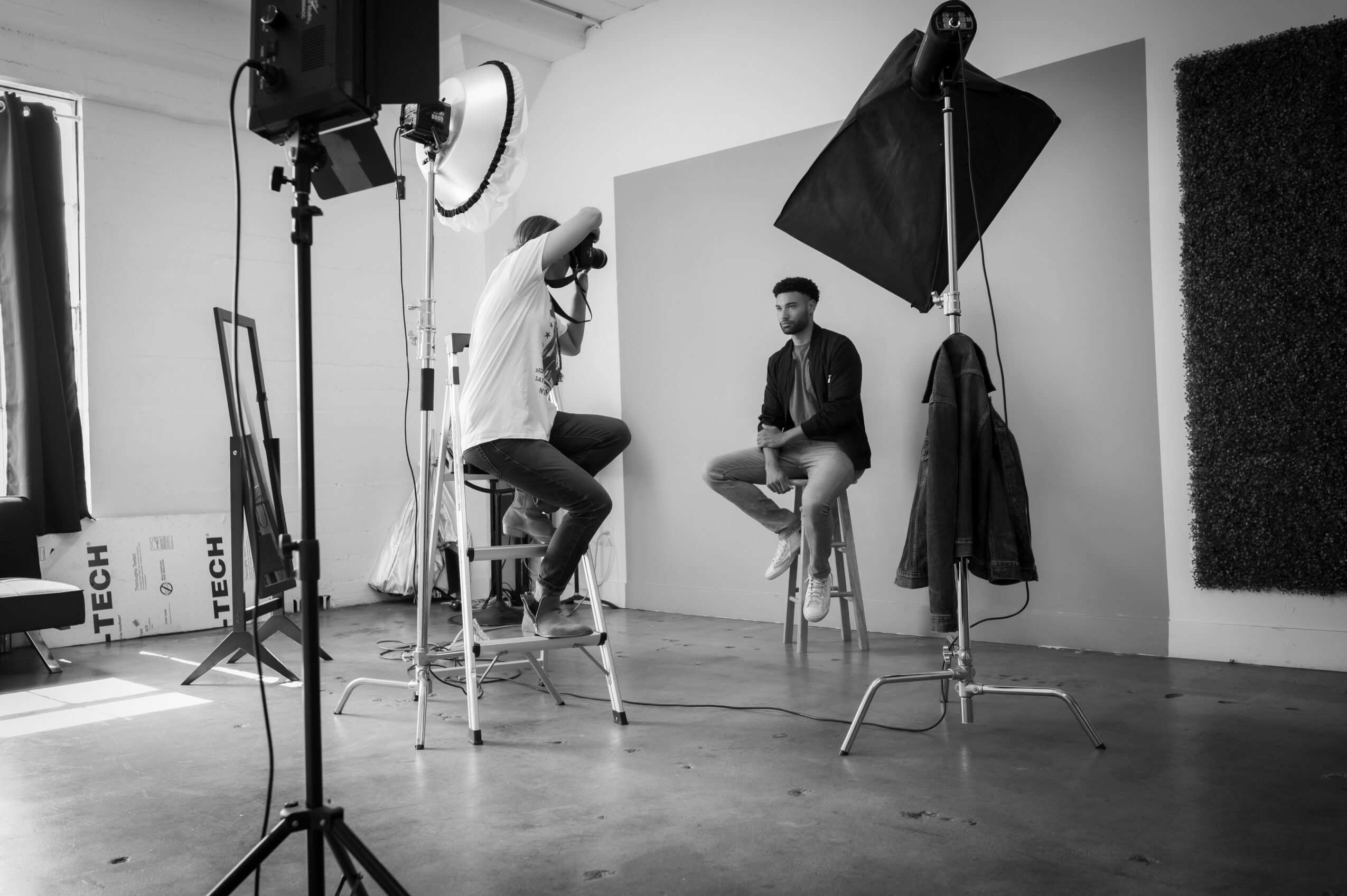 Two men sit on stools in a photography studio with lighting equipment and a backdrop, one man is being photographed.