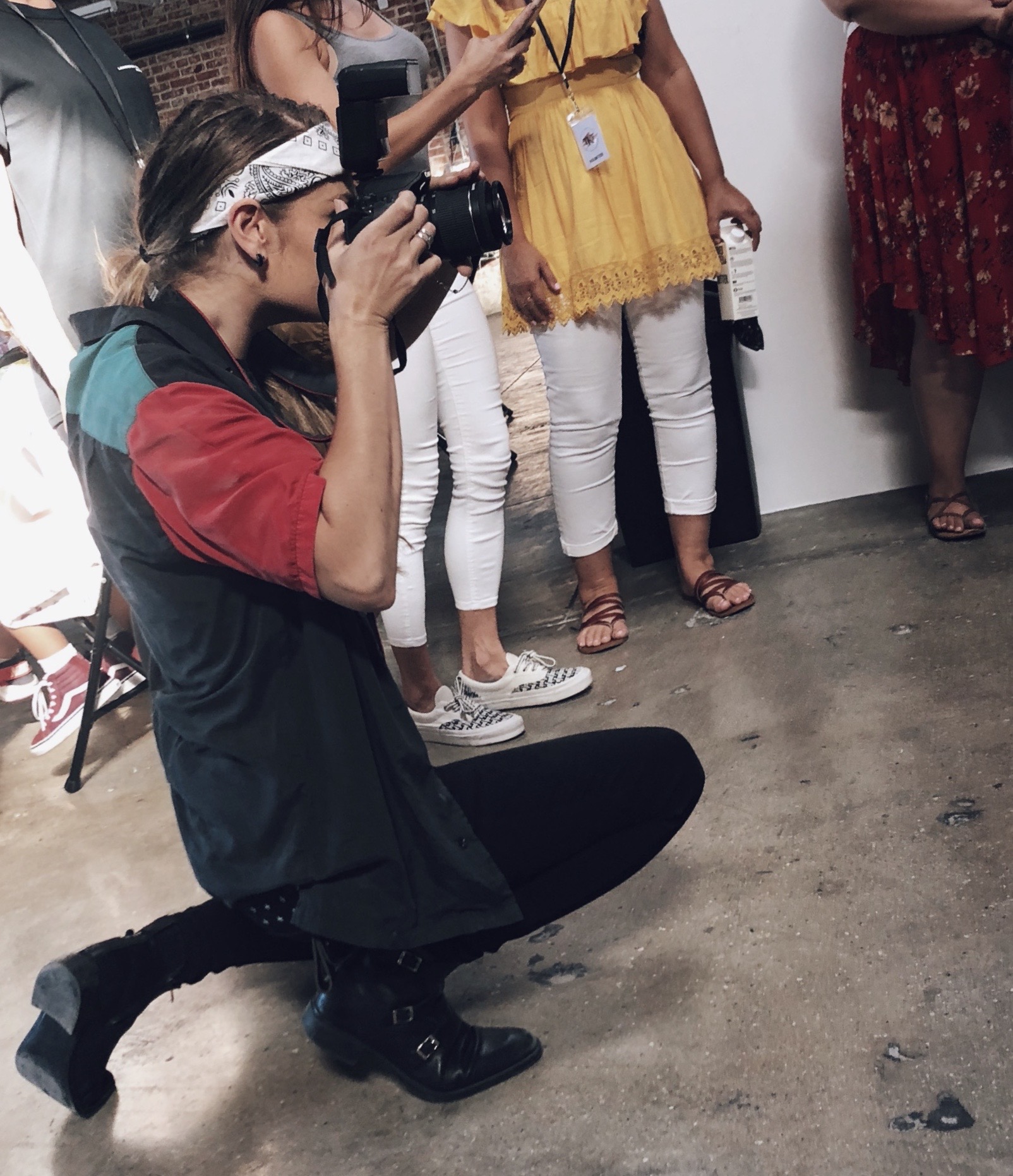 Person crouching with a camera, surrounded by people in colorful clothing, standing indoors on a concrete floor.