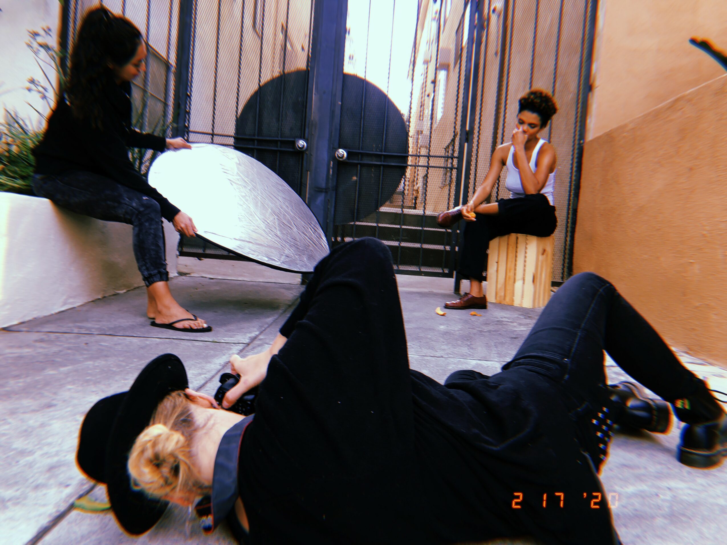 Three women in an indoor space, one lying on the floor with a hat, two sitting on a ledge, one holding a reflector.