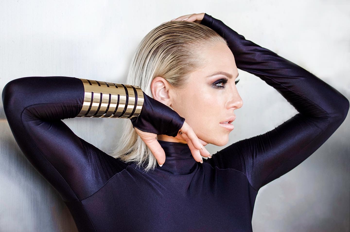 Woman with platinum blonde hair in a black outfit with gold and black arm accessories, posed with arms raised, looking sideways.