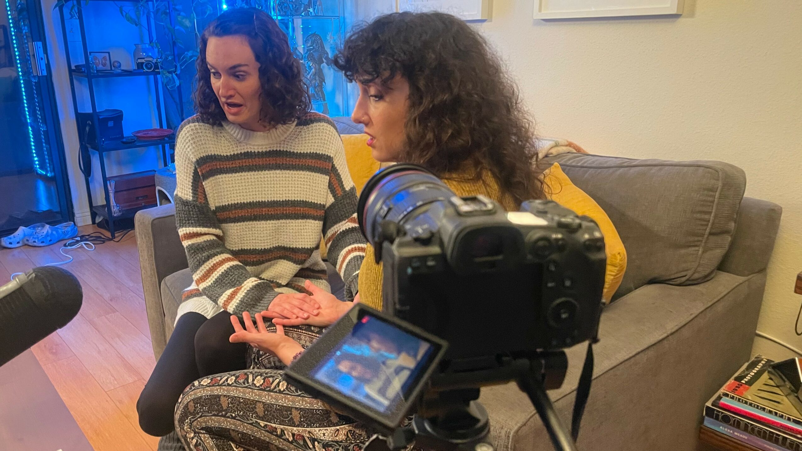 Two women sit on a sofa, one holding a smartphone, with a camera recording them in a room.