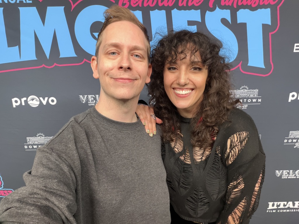 Two people smiling at an event with a backdrop featuring logos and text.