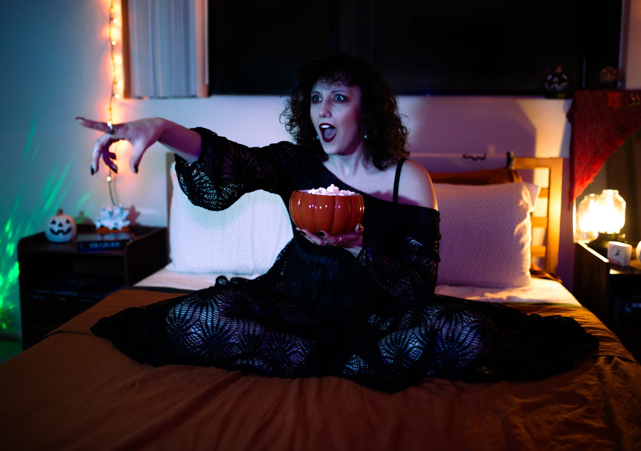 Person with curly hair sitting on a bed holding a pumpkin-shaped container, with Halloween decorations in the background.