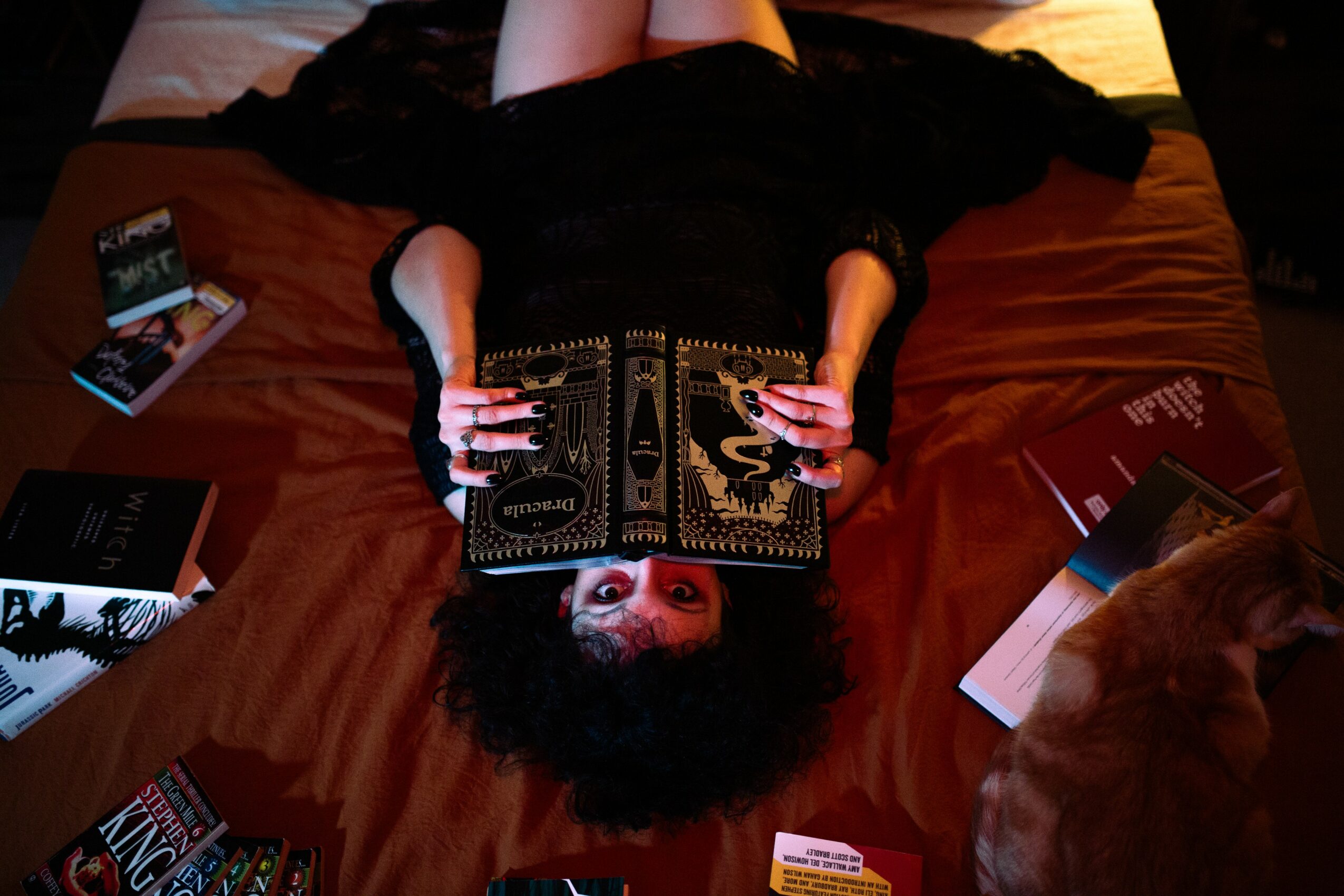 Person lying on bed reading a book, with a cat nearby, surrounded by books and magazines.