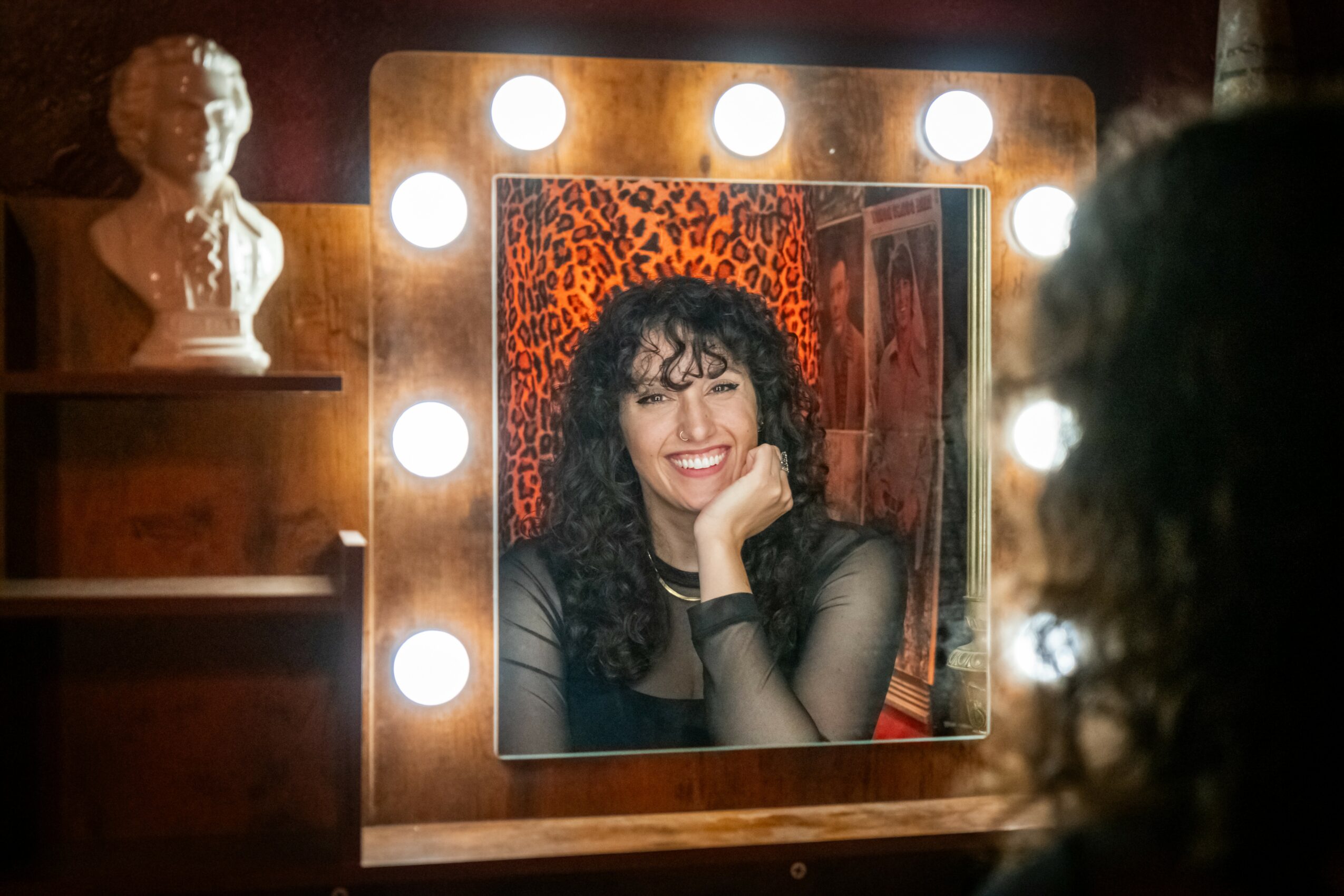 Woman with curly hair smiling at mirror with lights, in a room with a bust sculpture and leopard print background.