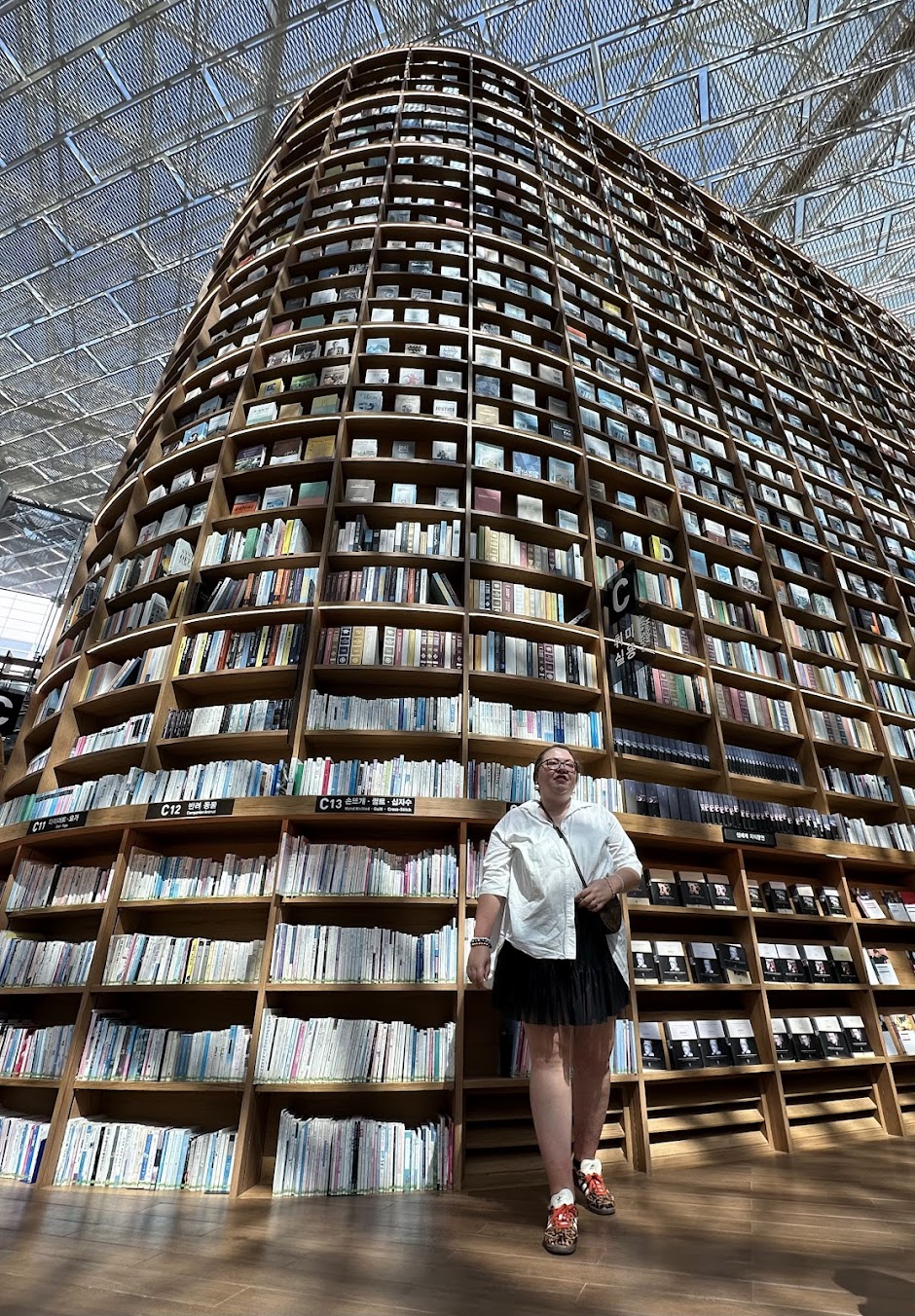 Person standing in front of a large, curved bookshelf filled with books inside a modern building with a glass ceiling.
