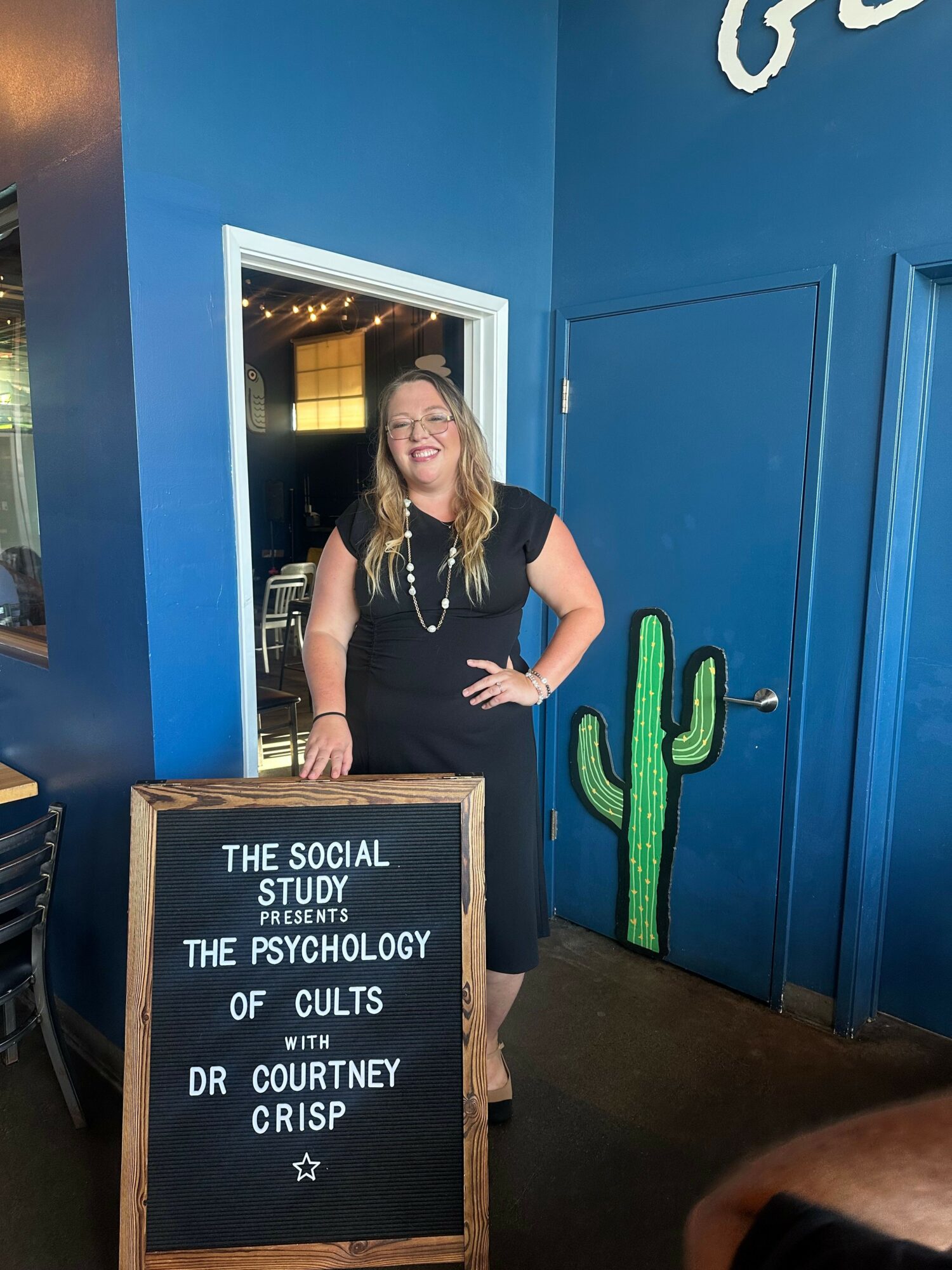 Woman standing in doorway with blue wall and cactus decoration, holding a drink, next to a blackboard sign, in an indoor setting.