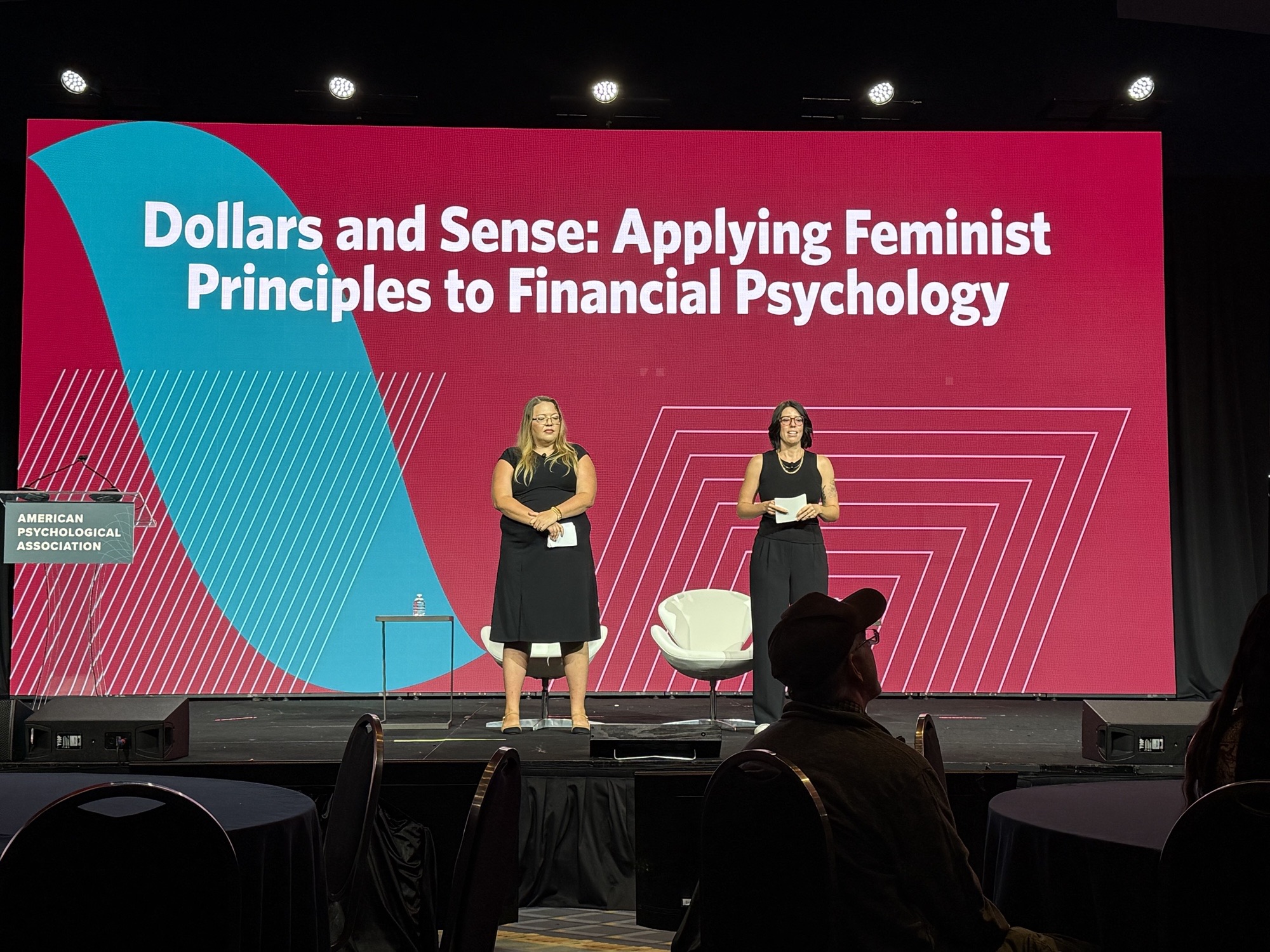 Two women stand on stage in front of a large pink and blue presentation screen with text about applying feminist principles to financial psychology.