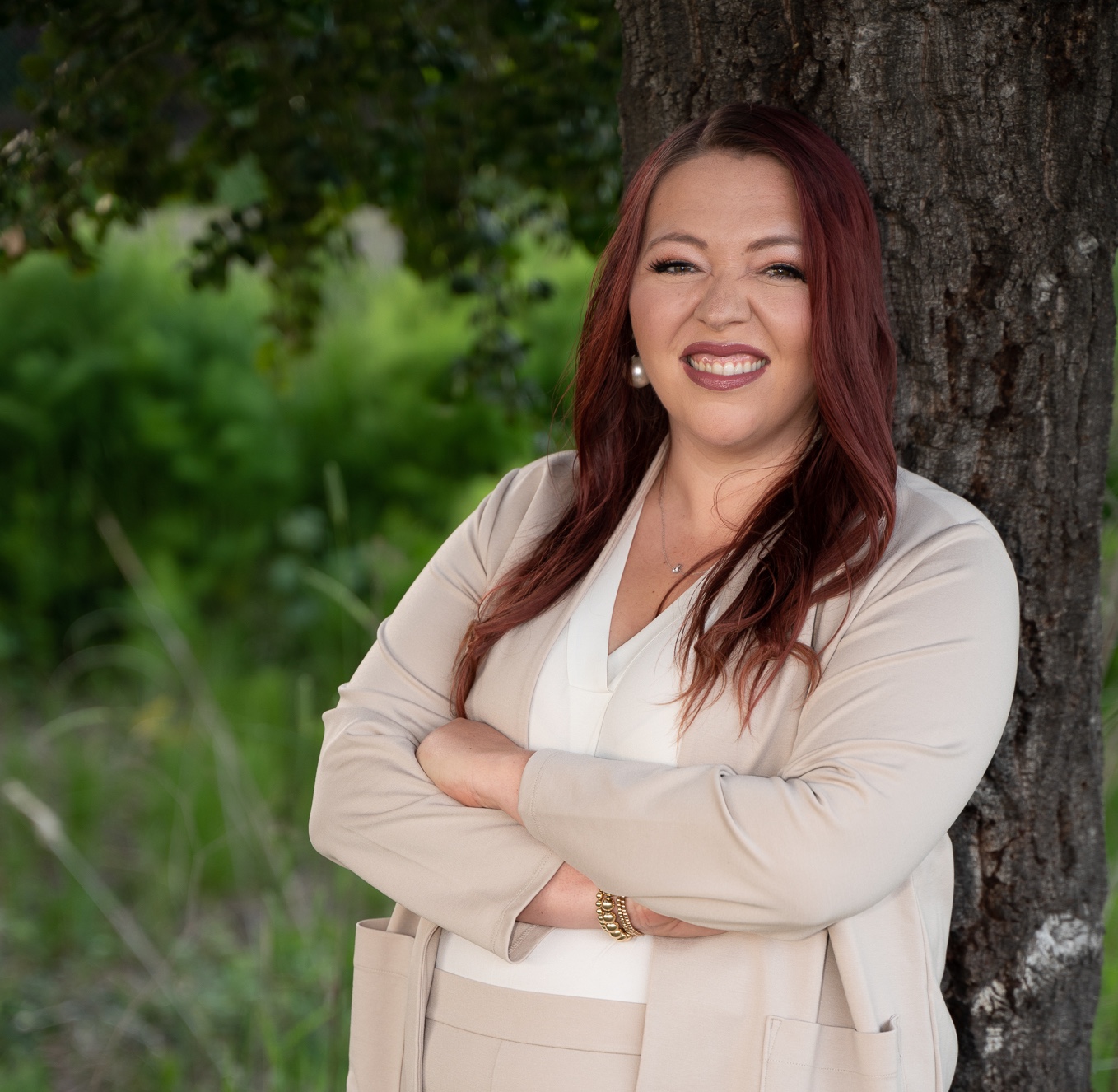Woman with long red hair leaning against a tree outdoors, smiling, with green foliage in background.