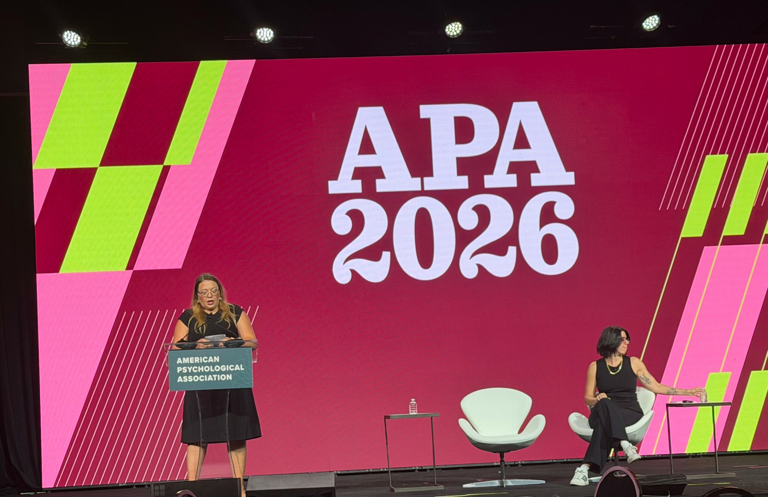 Two women on stage with a large pink background displaying 'APA 2026' and colorful geometric patterns.