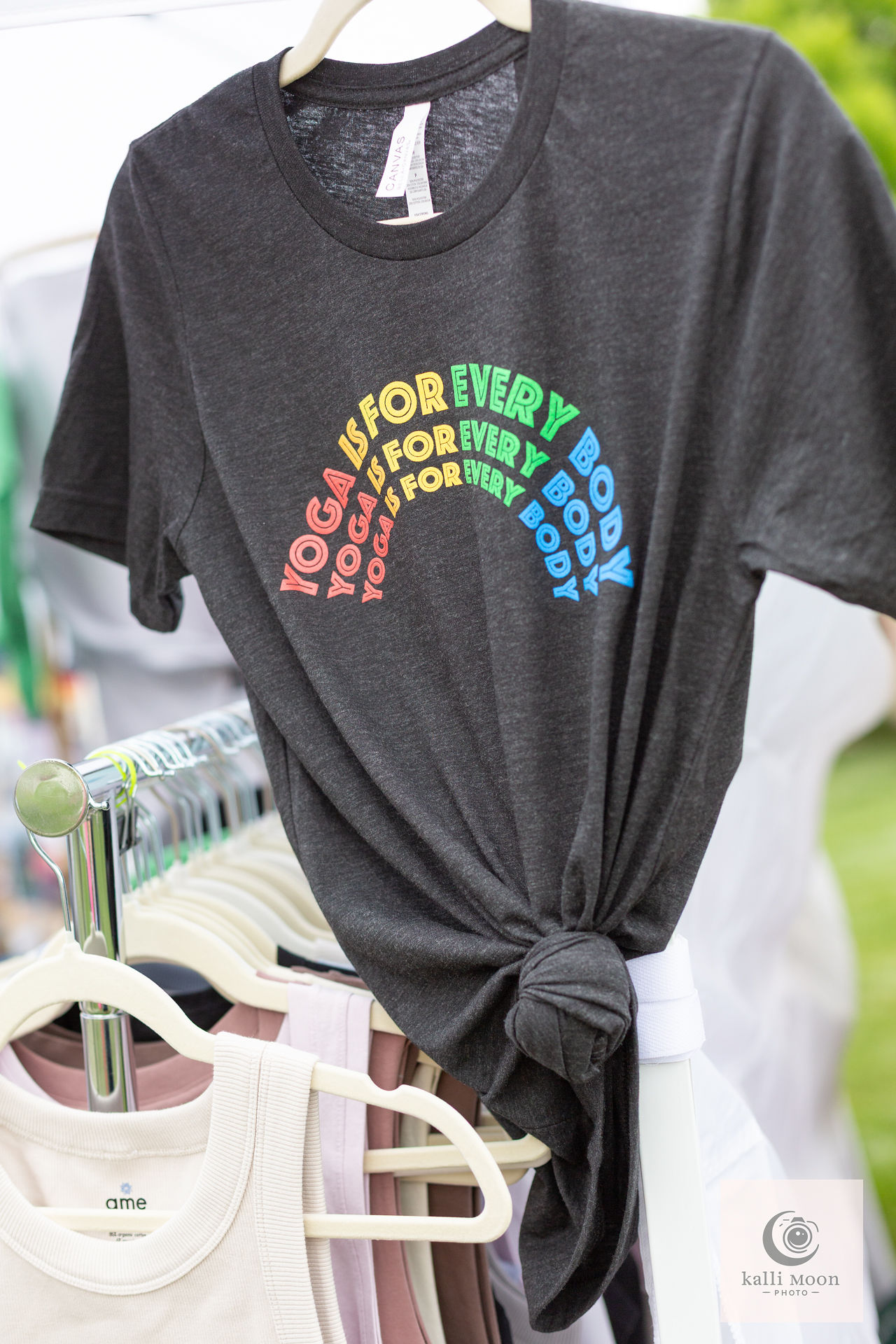 Black T-shirt with colorful rainbow text, tied in a knot at the waist, hanging on a hanger outdoors.