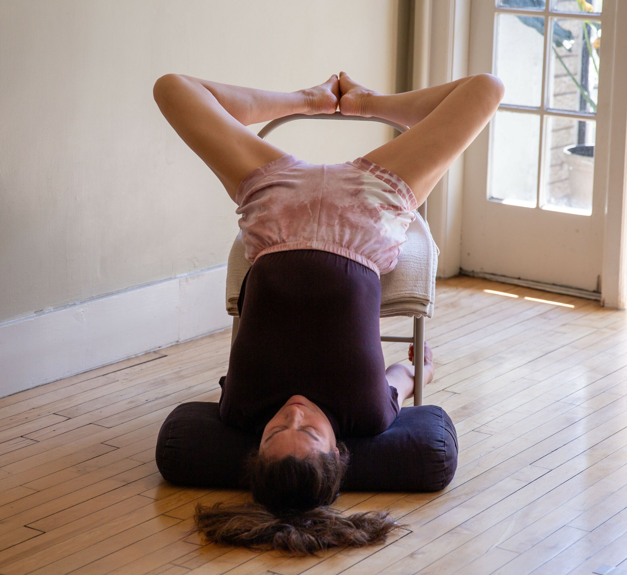 Person lying on a chair with legs over head, head and shoulders on the floor, in a room with wooden floor and door with window.