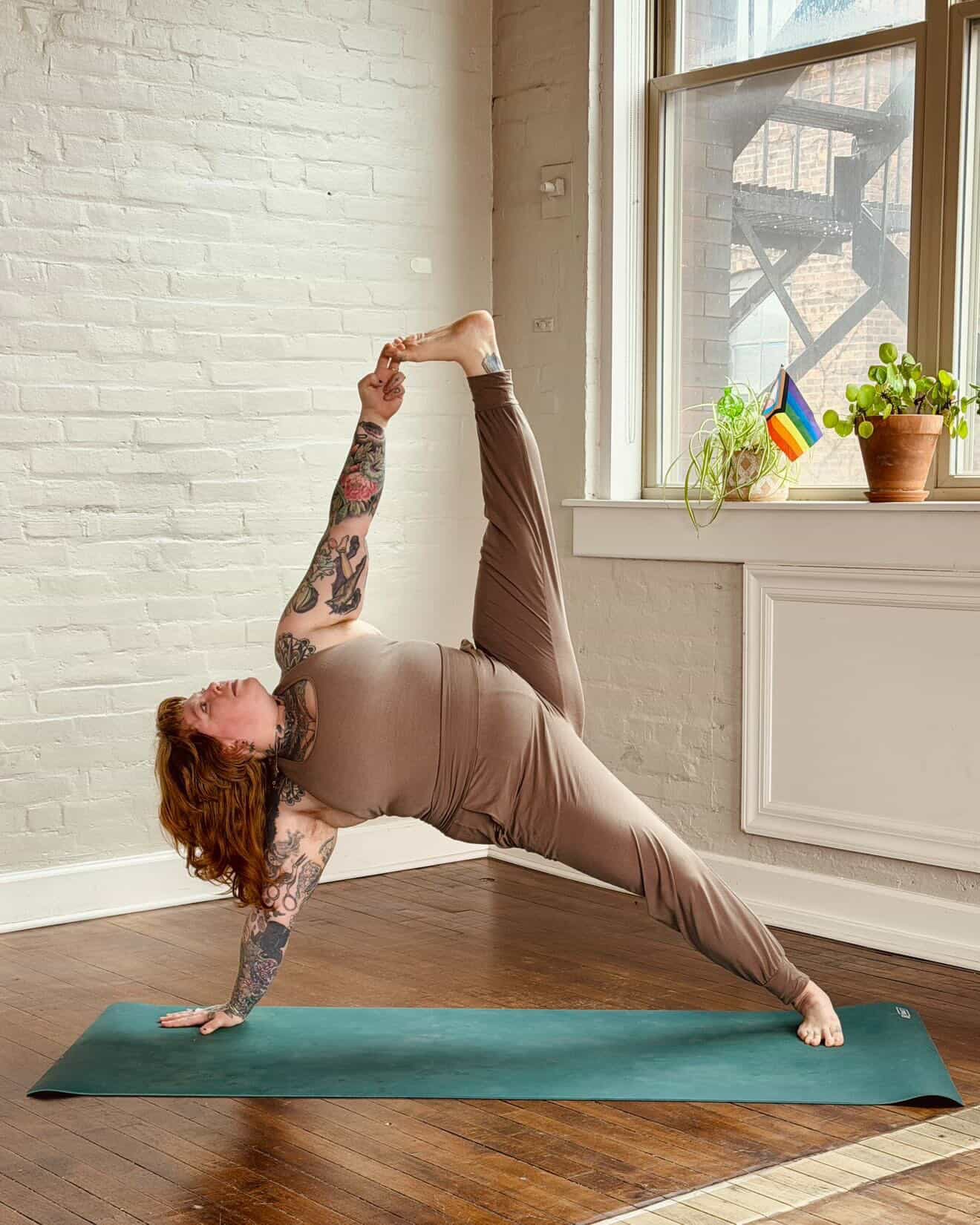 Woman practicing yoga in a room with large window, white brick wall, and potted plants, holding foot behind her head in a side stretch pose.